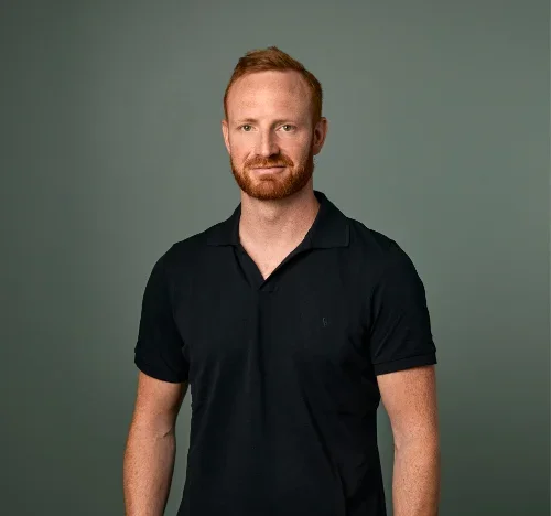 En mann med rødaktig hår og skjegg, iført svart polo, står mot en grågrønn bakgrunn.