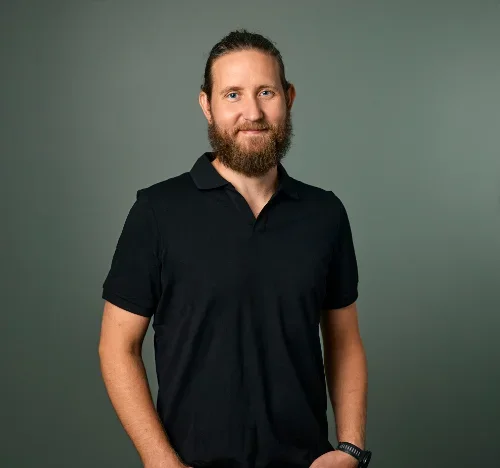 En mann med langt hår og skjegg, iført svart T-skjorte, står mot en grå bakgrunn og smiler.