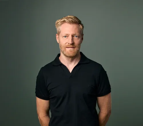 En mann med rød hår og skjegg, iført svart polo shirt, står foran en grå bakgrunn.