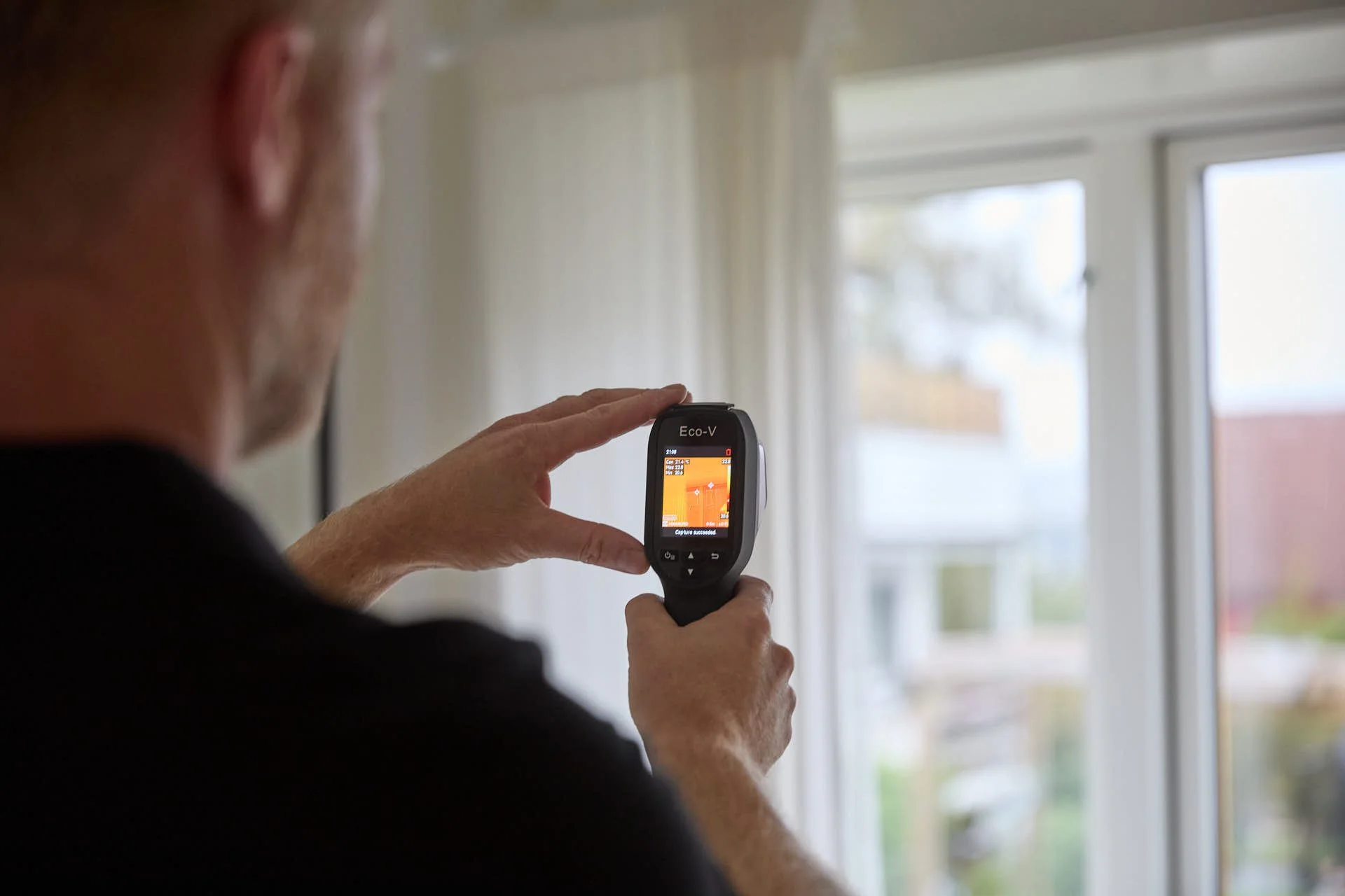 En person holder en digital termometer foran et stort vindu utendørs, med fokus på termometeret.