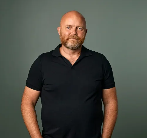 Portrett av en mann med skjegg i svart polo-skjorte mot en grå bakgrunn.