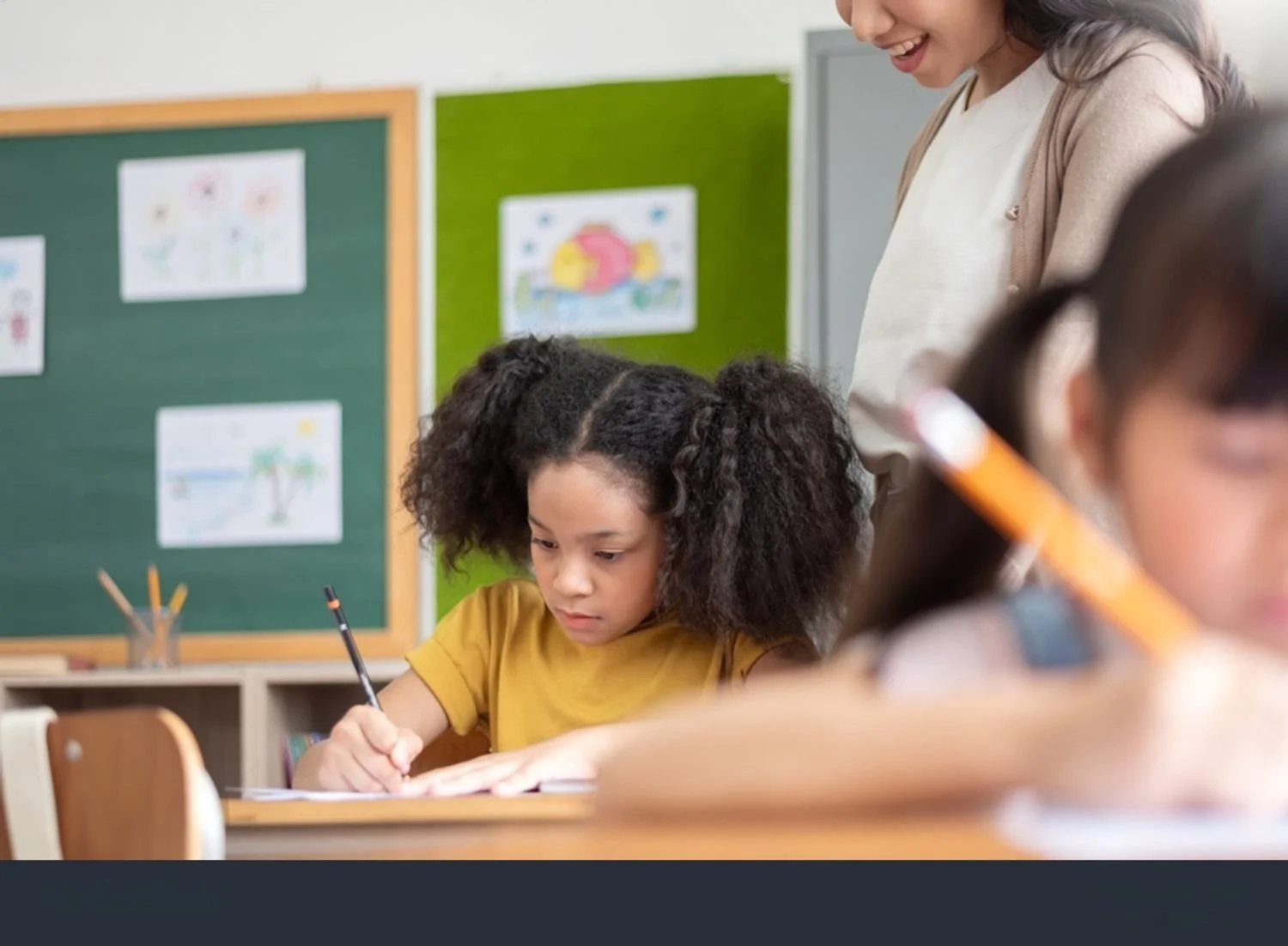 A young girl with curly hair writing in a notebook in a classroom, with a teacher standing over her, watching. The classroom has a chalkboard with drawings on it.