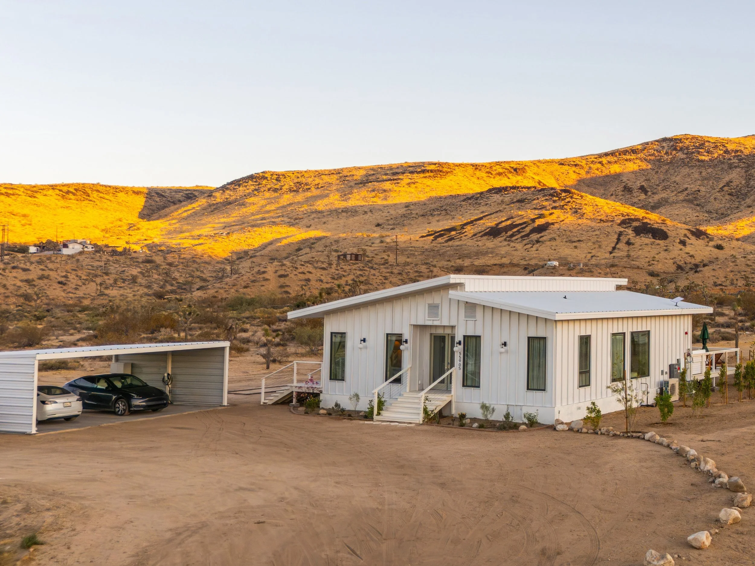 A white modern house with a small porch, surrounded by a desert landscape with rolling hills in the background, at sunset.