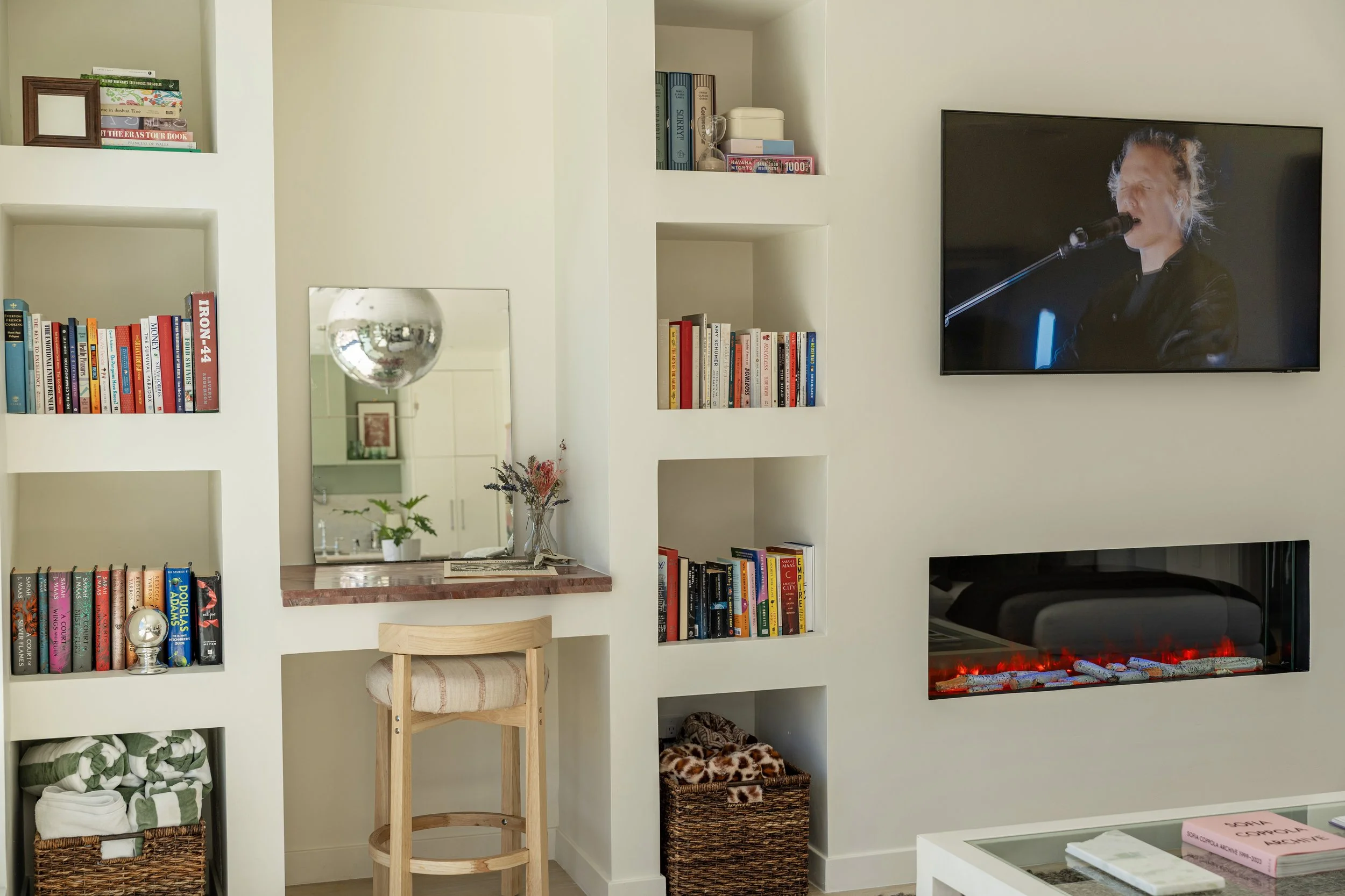 Living room with built-in white shelving filled with books and decorative items, a wall-mounted TV showing a singer, a stylish electric fireplace, a wooden bar stool, a mirror, and assorted baskets and decor items.