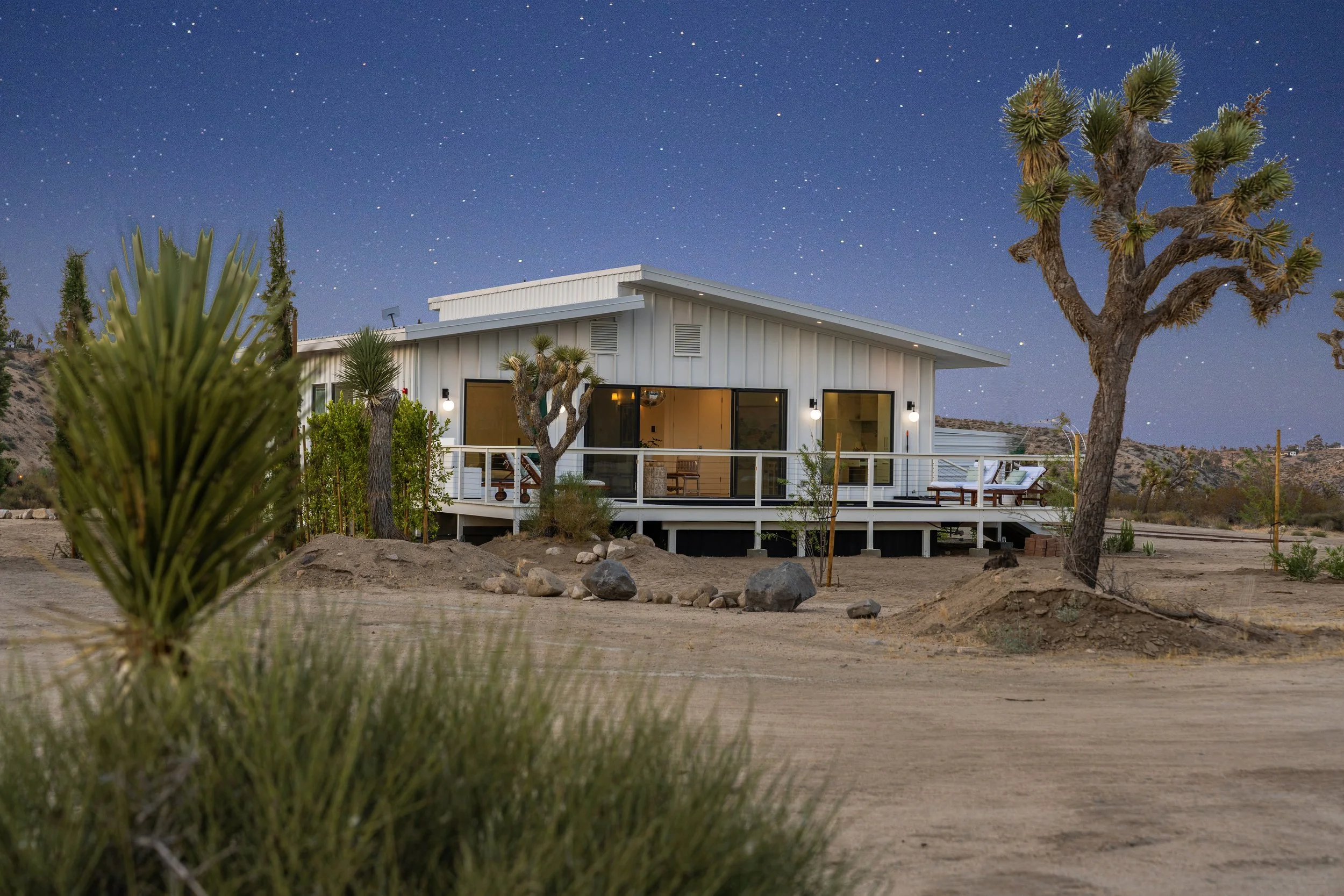 A modern house with large glass doors and windows, surrounded by desert landscape featuring Joshua trees, rocks, and sandy terrain, under a starry night sky.