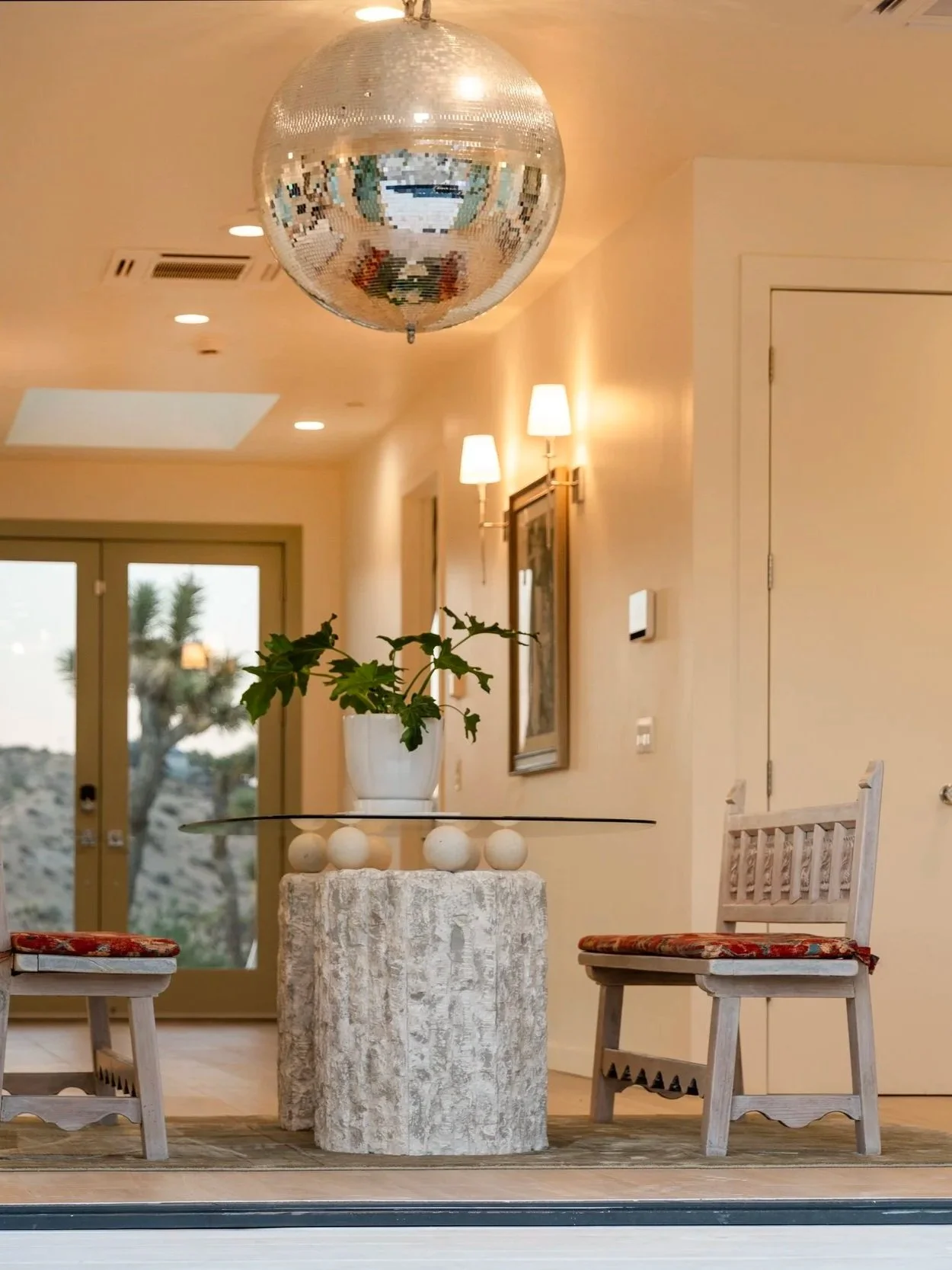 Interior of a living space with a disco ball, a round stone table, two white wooden chairs with red cushions, a plant in a white pot, and warm wall lighting.