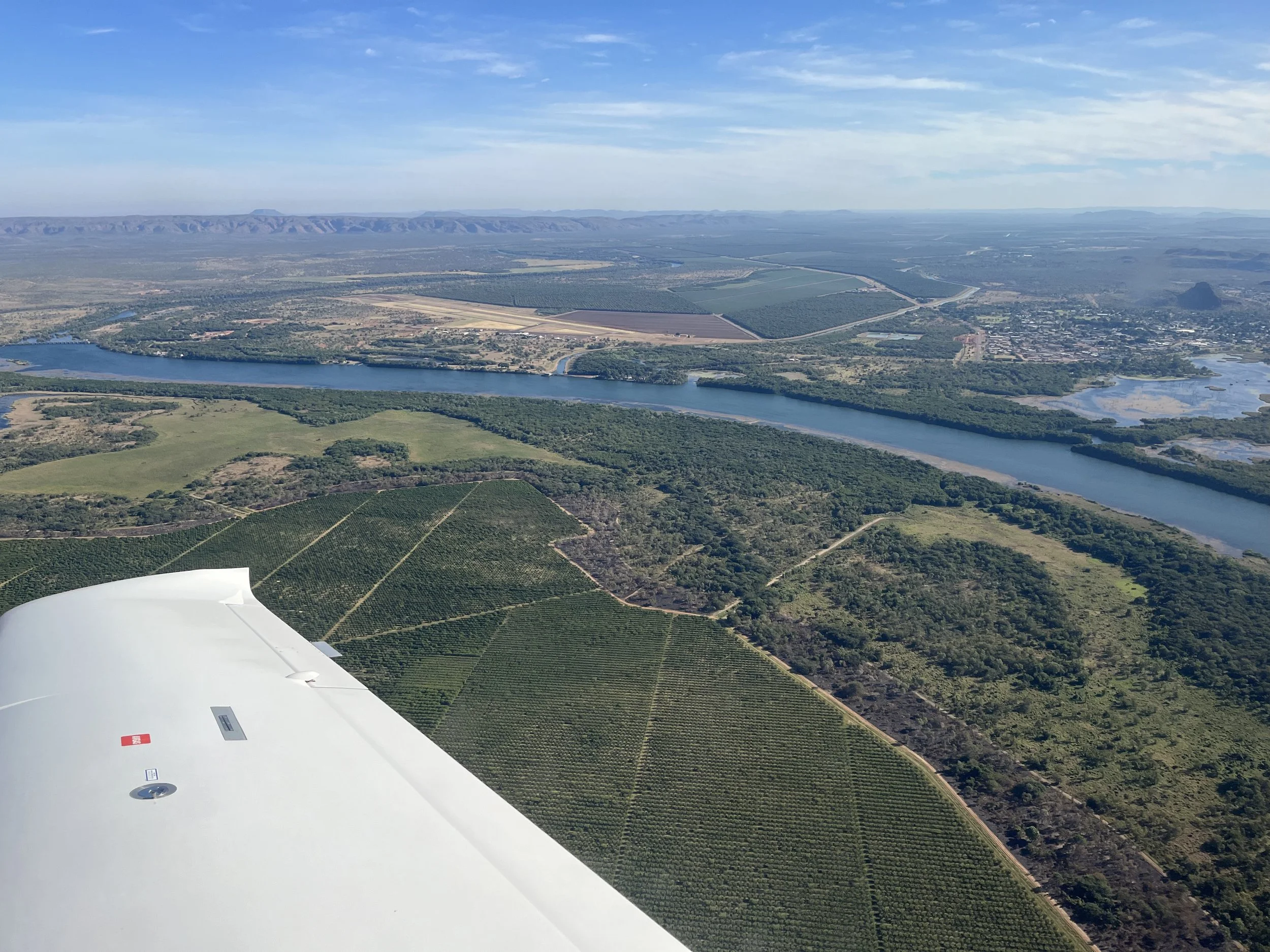 Aerial view of a river flowing through green fields, farmland, and a small town, with mountains in the distance, taken from an airplane.