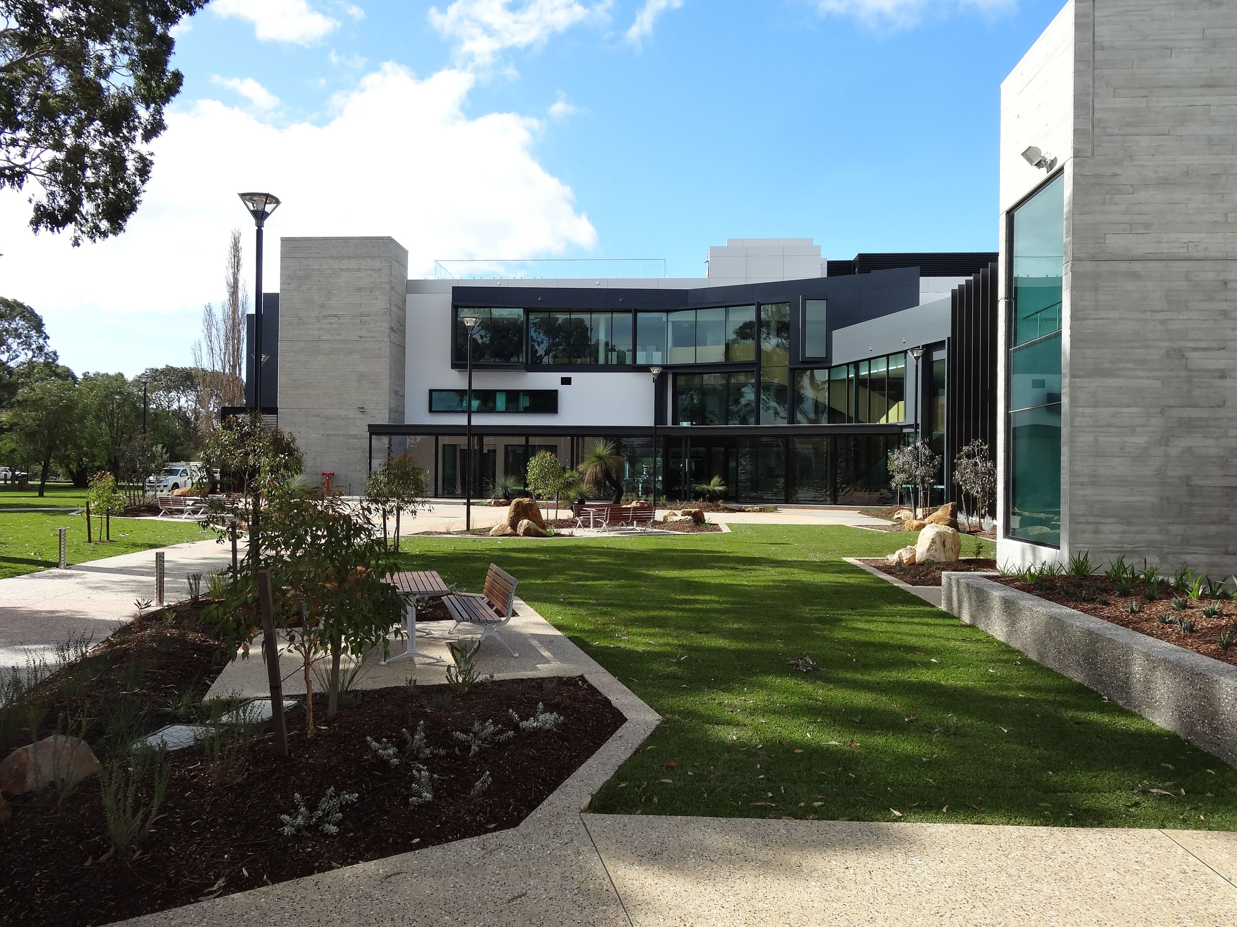 Modern building with large glass windows, landscaped courtyard with benches, trees, and grass, under a partly cloudy sky.