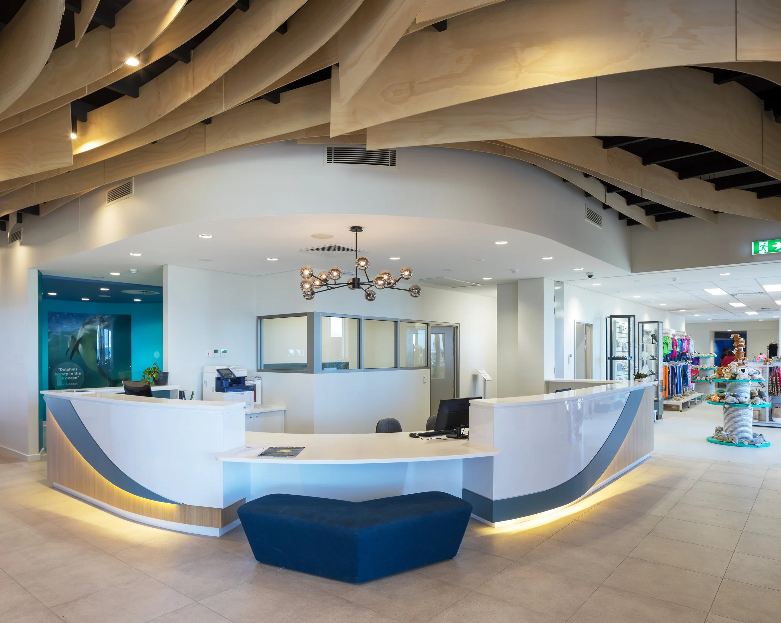 Modern hotel reception area with a curved white check-in desk, black accents, and a unique lighting fixture hanging from the ceiling. To the right, there are shelves with store merchandise including toys and clothing. The ceiling features wooden pane