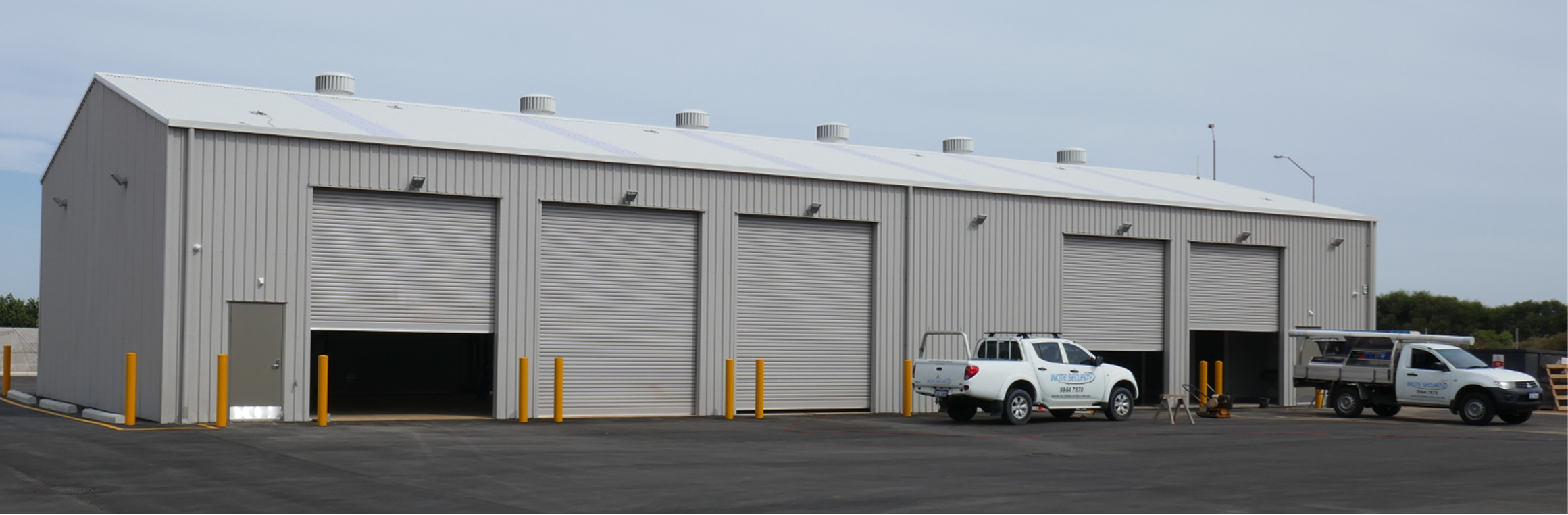 A large industrial warehouse with four rolled-up garage doors, two trucks parked outside, and a mostly cloudy sky overhead.