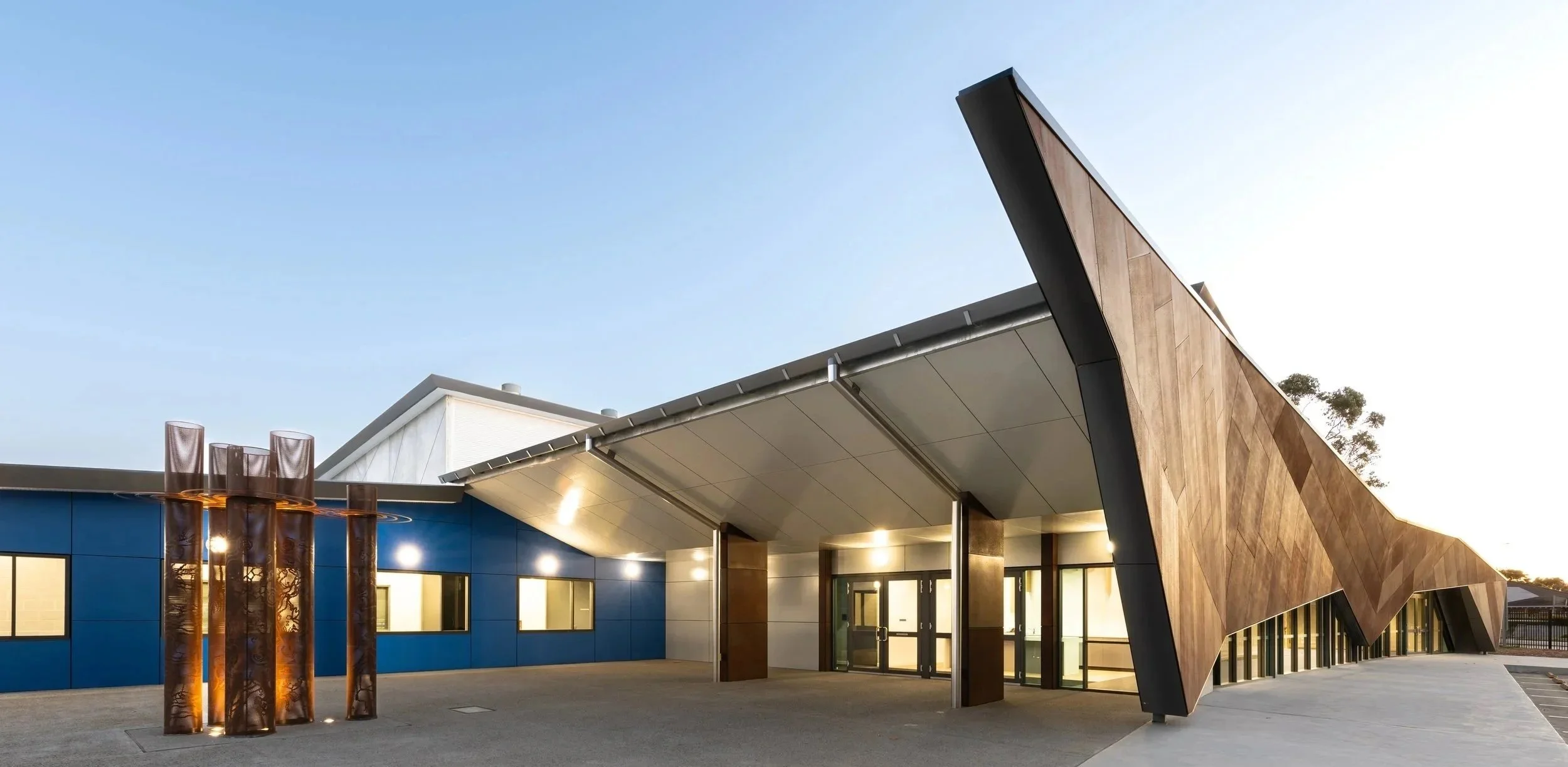 Modern building with a distinctive angular wooden facade, blue wall, and decorative copper-like sculptures outside, under a clear sky.