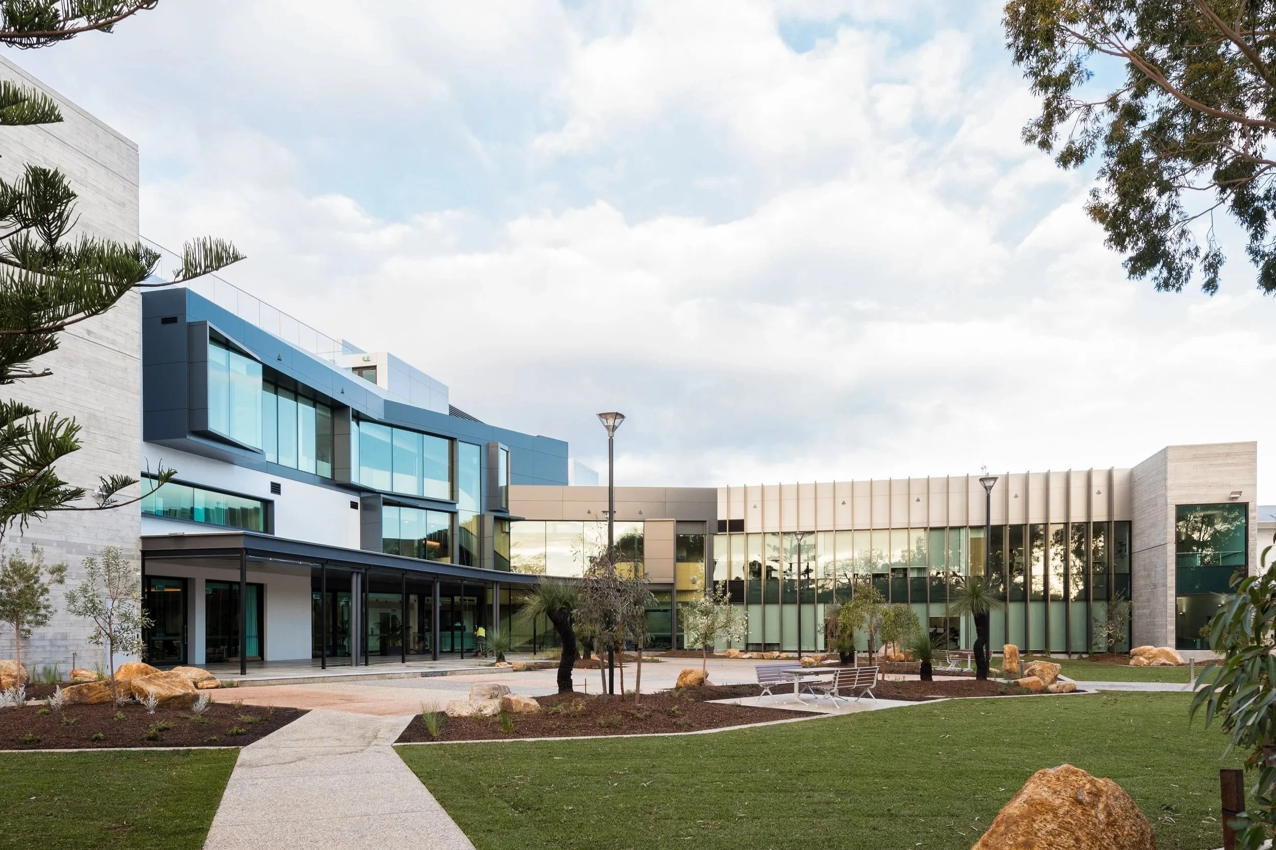 Modern building with large glass windows, landscaped courtyard with trees, rocks, benches, and pathways, under a partly cloudy sky.