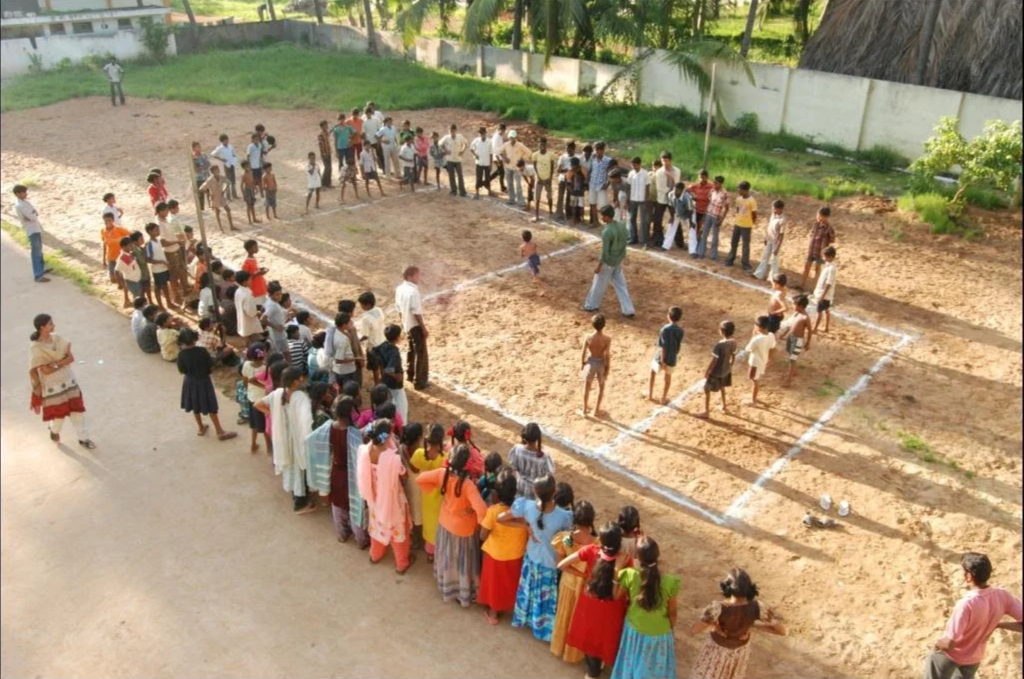 Children playing volleyball on a dirt court while spectators watch, outdoors.