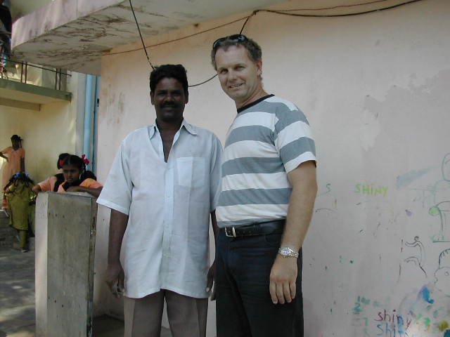Two men standing side by side outdoors, smiling at the camera, with a group of children in the background near a wall with colorful drawings.