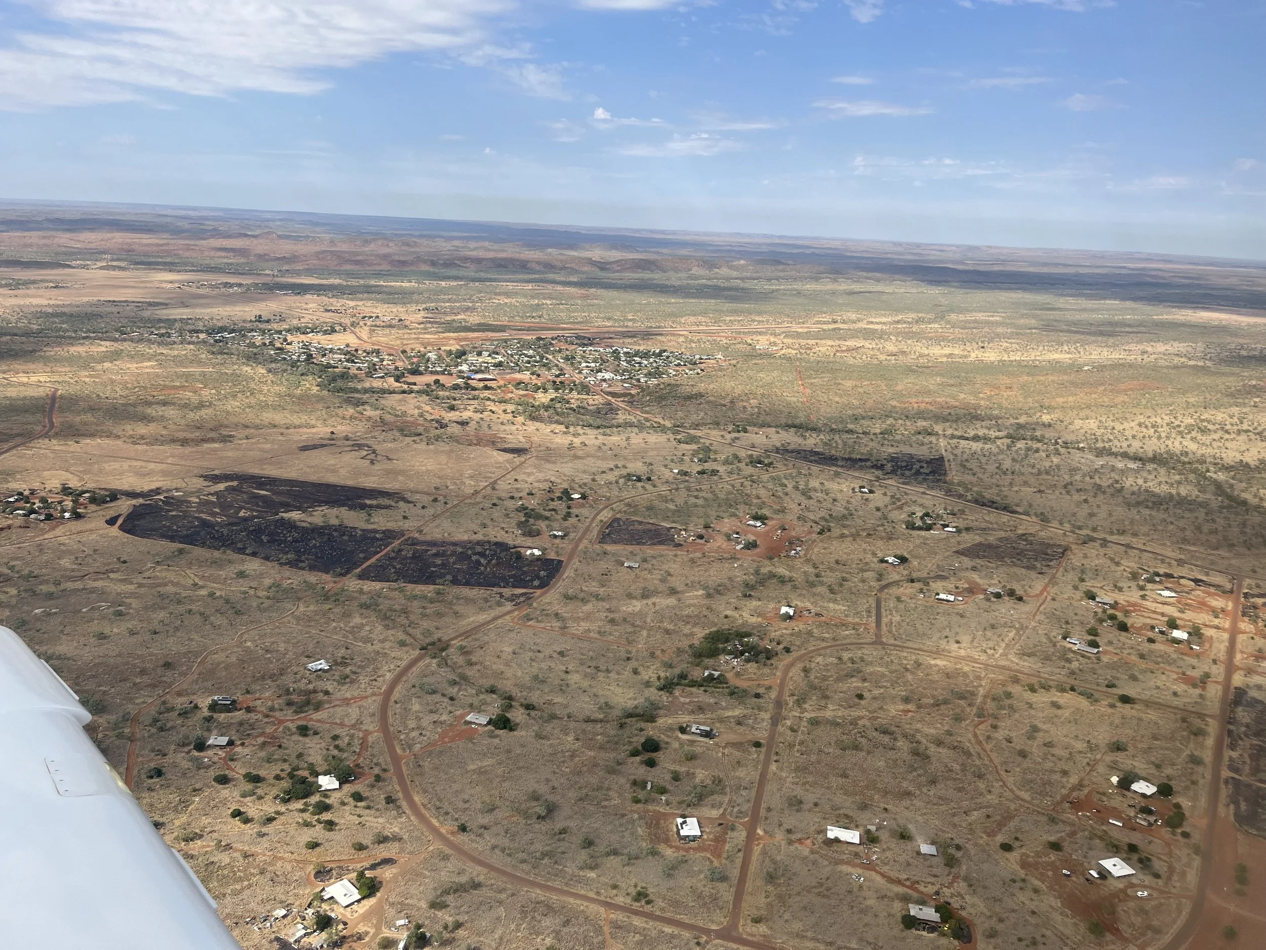 Aerial view of a semi-arid landscape with scattered houses, dirt roads, and patches of blackened land from recent fires, under partly cloudy skies.