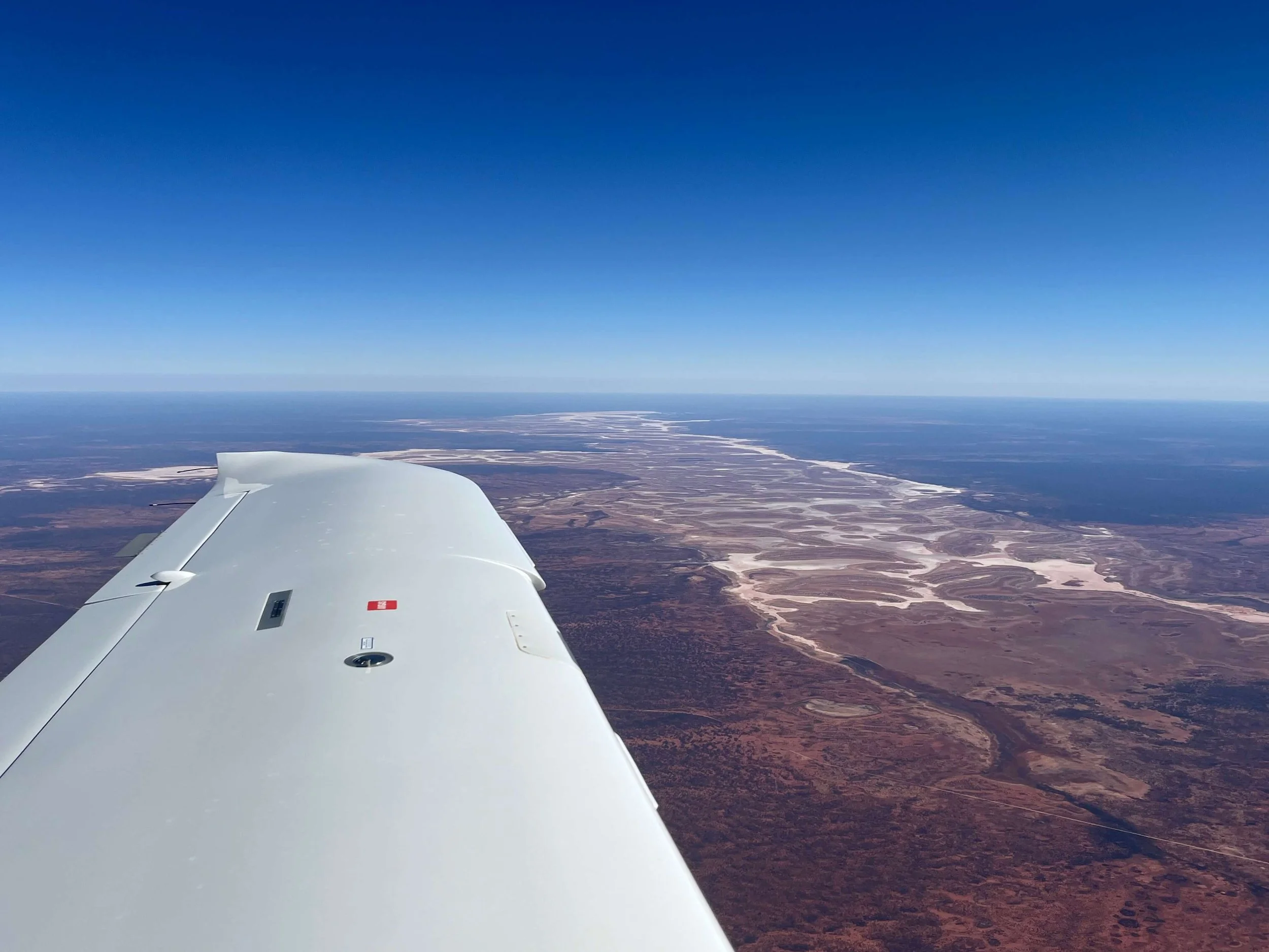View from an airplane window showing a landscape of dry terrain and a winding river below a clear blue sky.