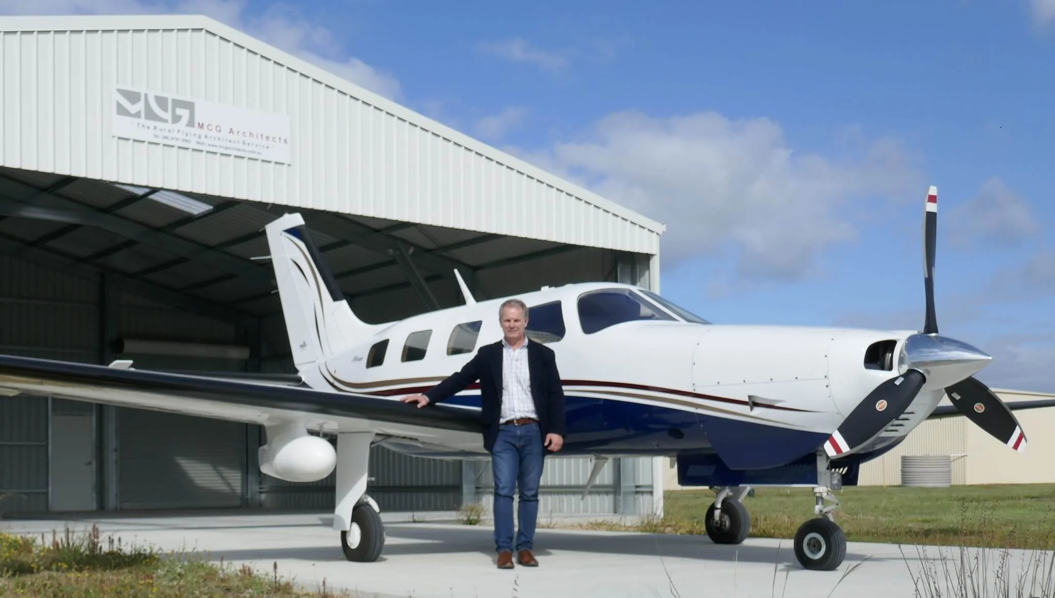 Man standing next to a small white and blue private airplane outside of a hangar on a sunny day.