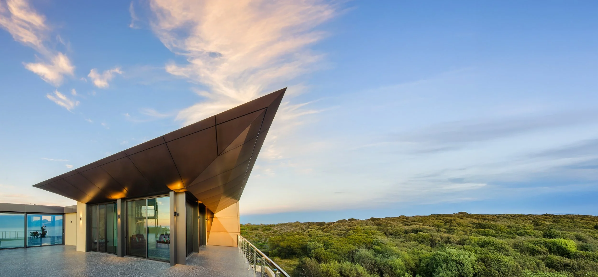 Modern building with an angular roof and large glass windows overlooking a lush green landscape and a blue sky with clouds.