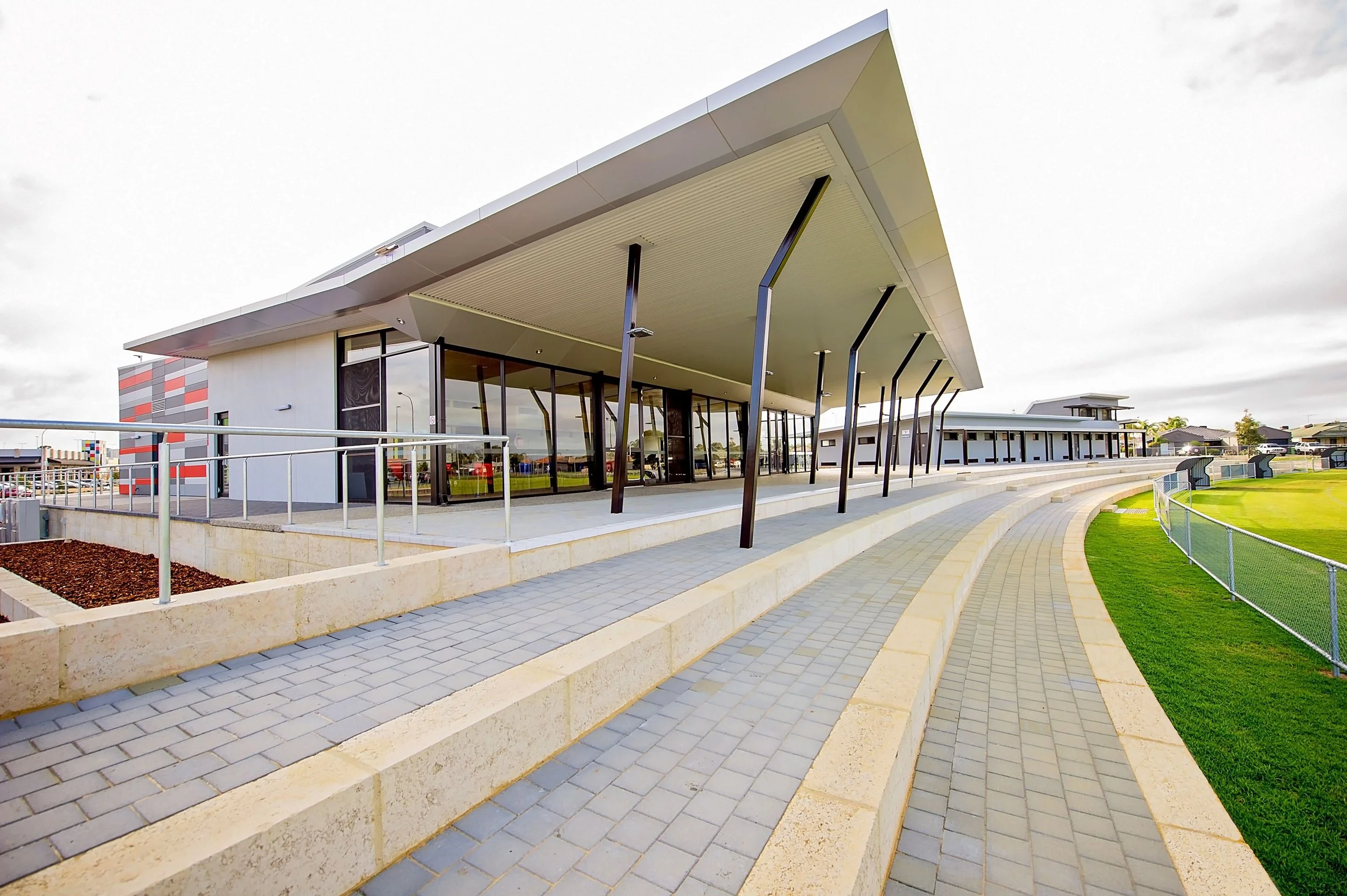 Modern building with large glass windows and sloped roof supported by angled metal columns, adjacent to a curved walkway with tiered seating, grassy field, and sports field fencing.