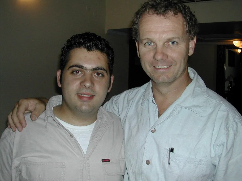 Two men standing close together smiling, one has his arm around the other's shoulder, in an indoor setting.