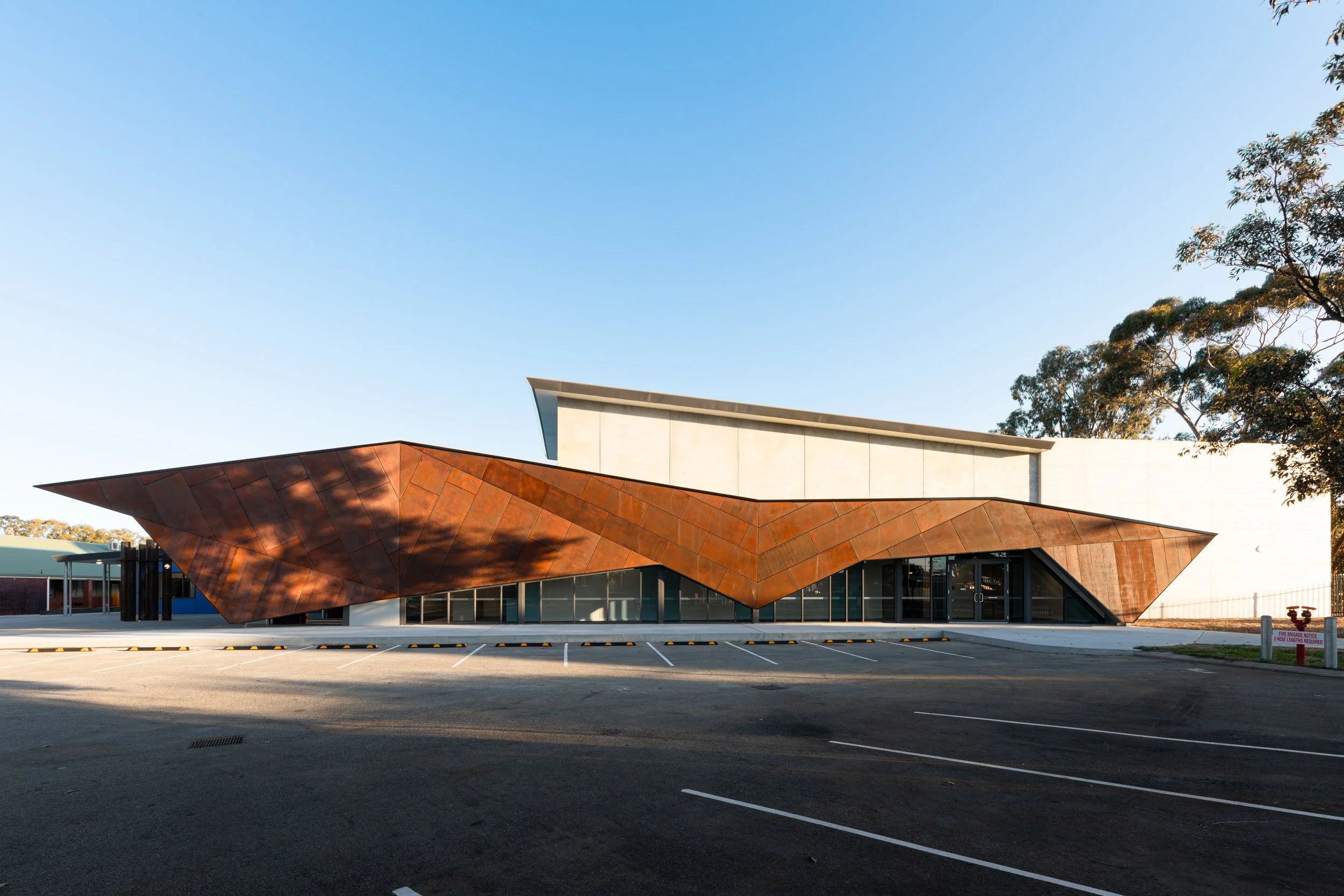 Modern building with a rust-colored, angular metal facade and large glass windows, with a clear blue sky and some trees in the background, and an empty parking lot in the foreground.