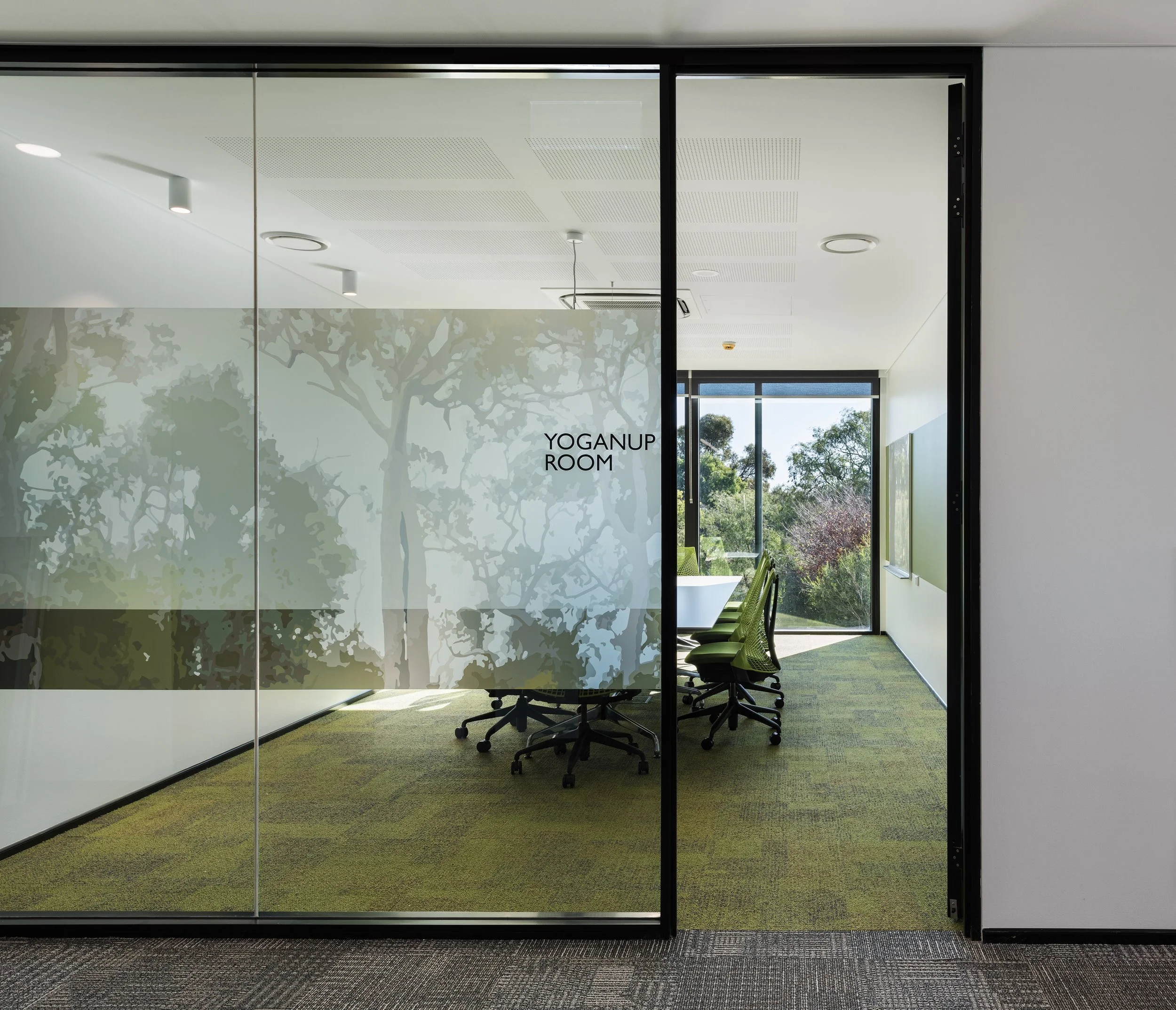 Glass door with frosted trees decal labeled 'Yoganup Room' leading to a conference room with green chairs and a large window showing trees outside.
