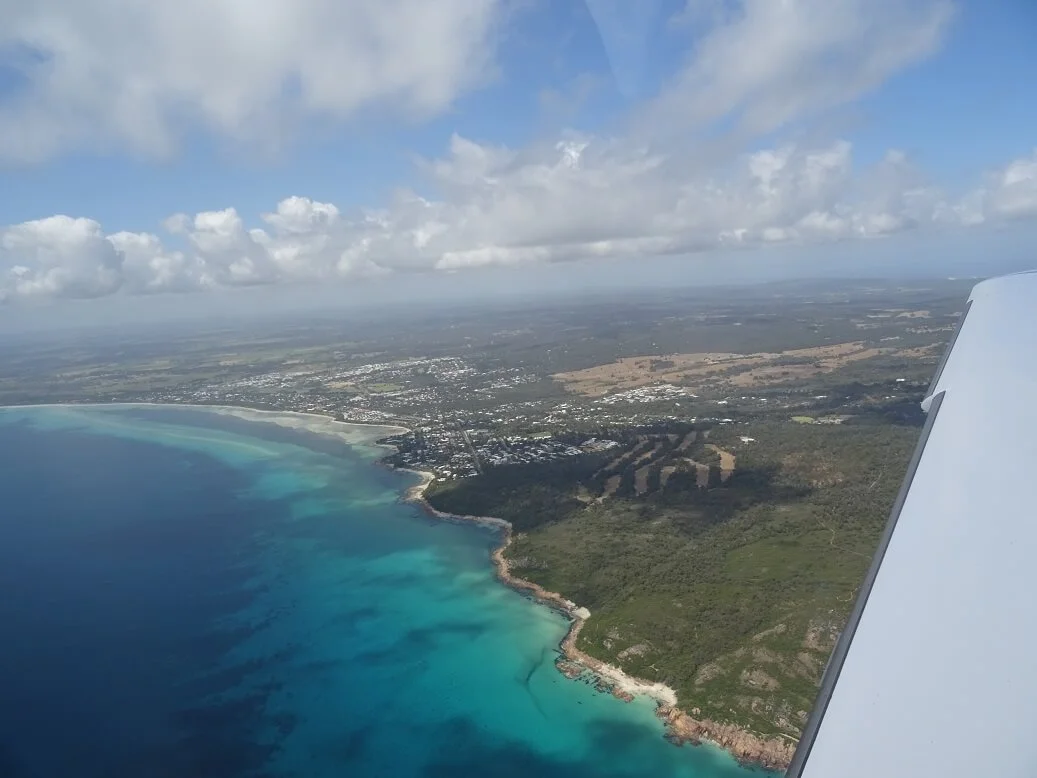 Aerial view of a coastline with turquoise water, sandy beaches, and green hills, with a small town or city in the distance. Part of an airplane wing is visible on the right side.
