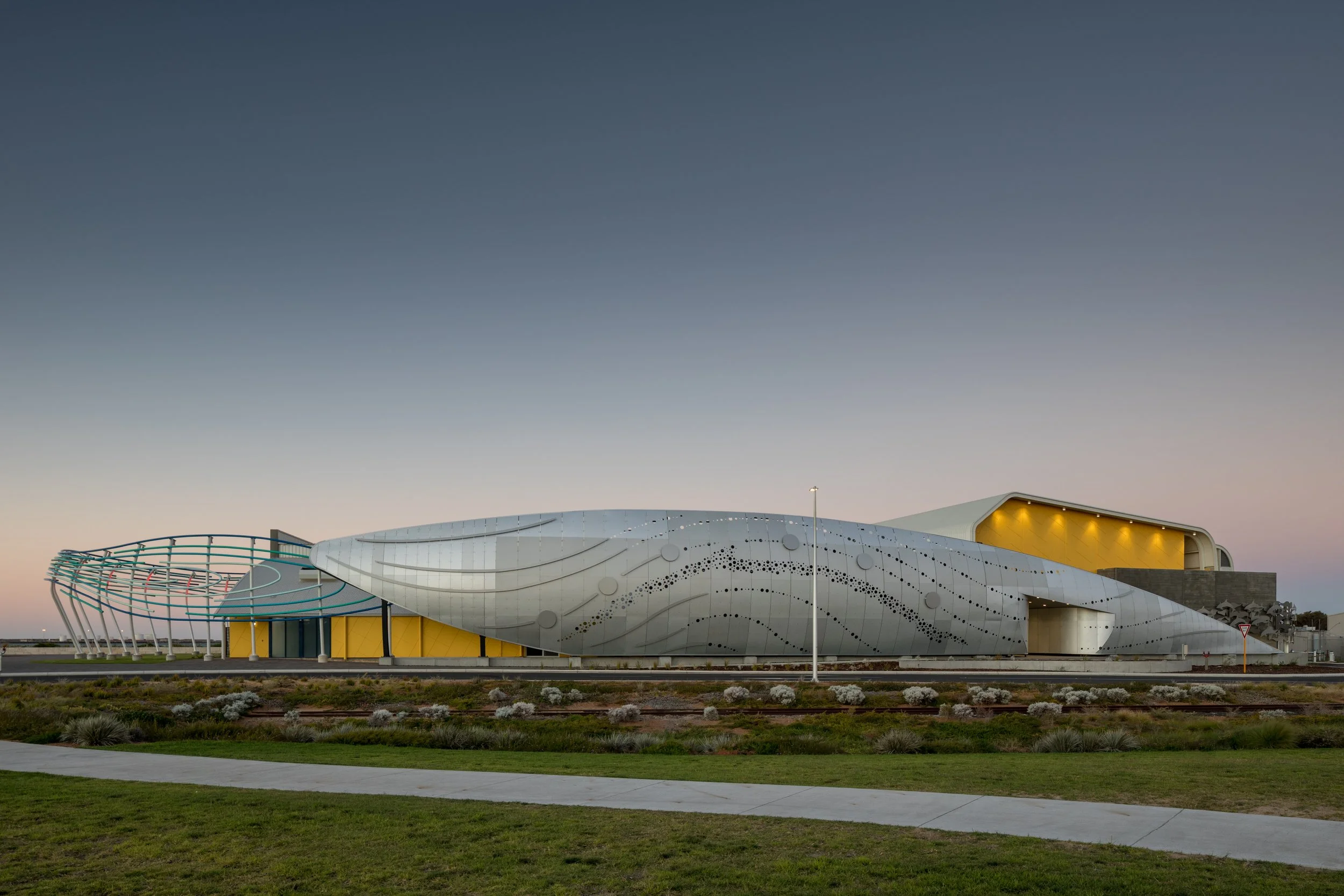 Modern building with curved, metallic exterior, large open windows, and decorative patterns, set against a twilight sky.