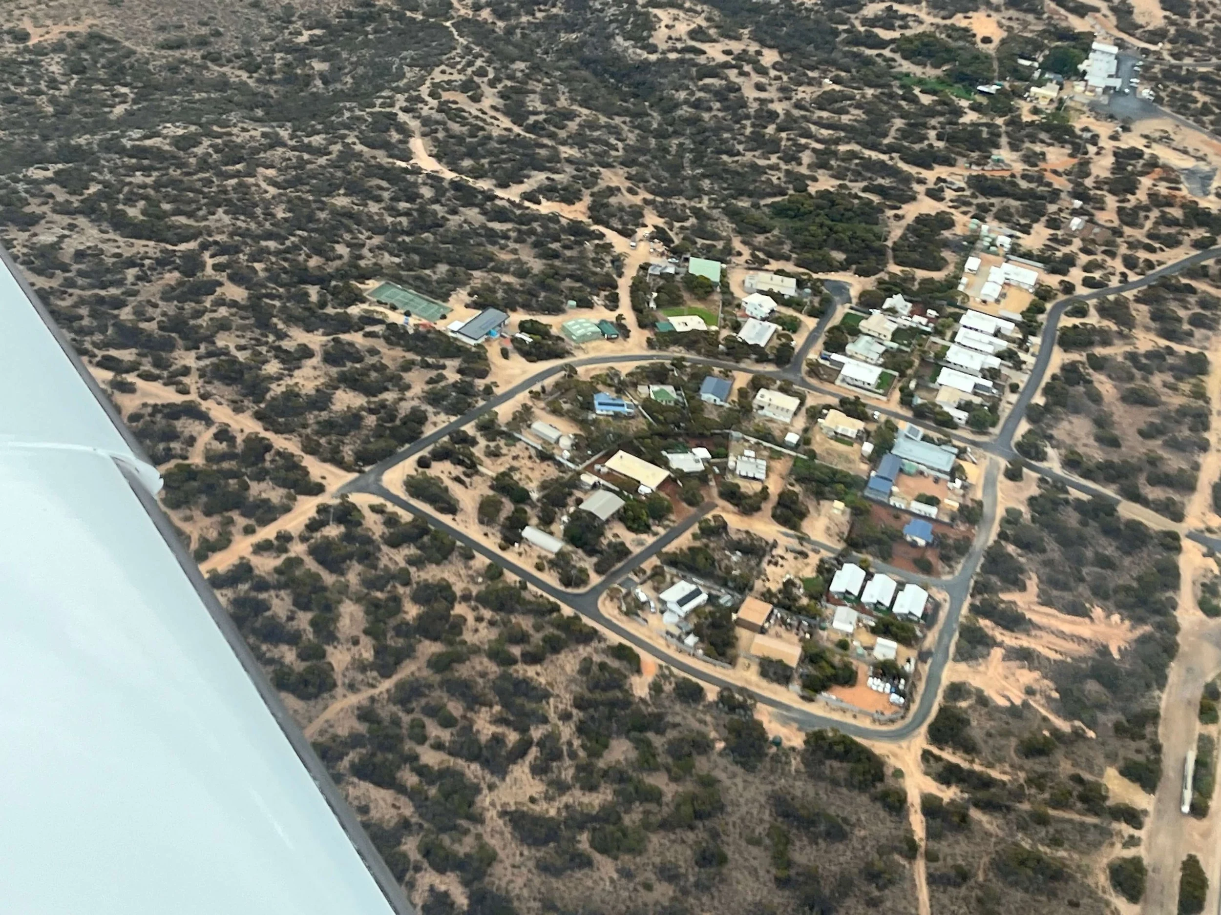 Aerial view of a Eucla, a small residential area with multiple houses, roads, and some green spaces, surrounded by a dry, desert-like landscape.