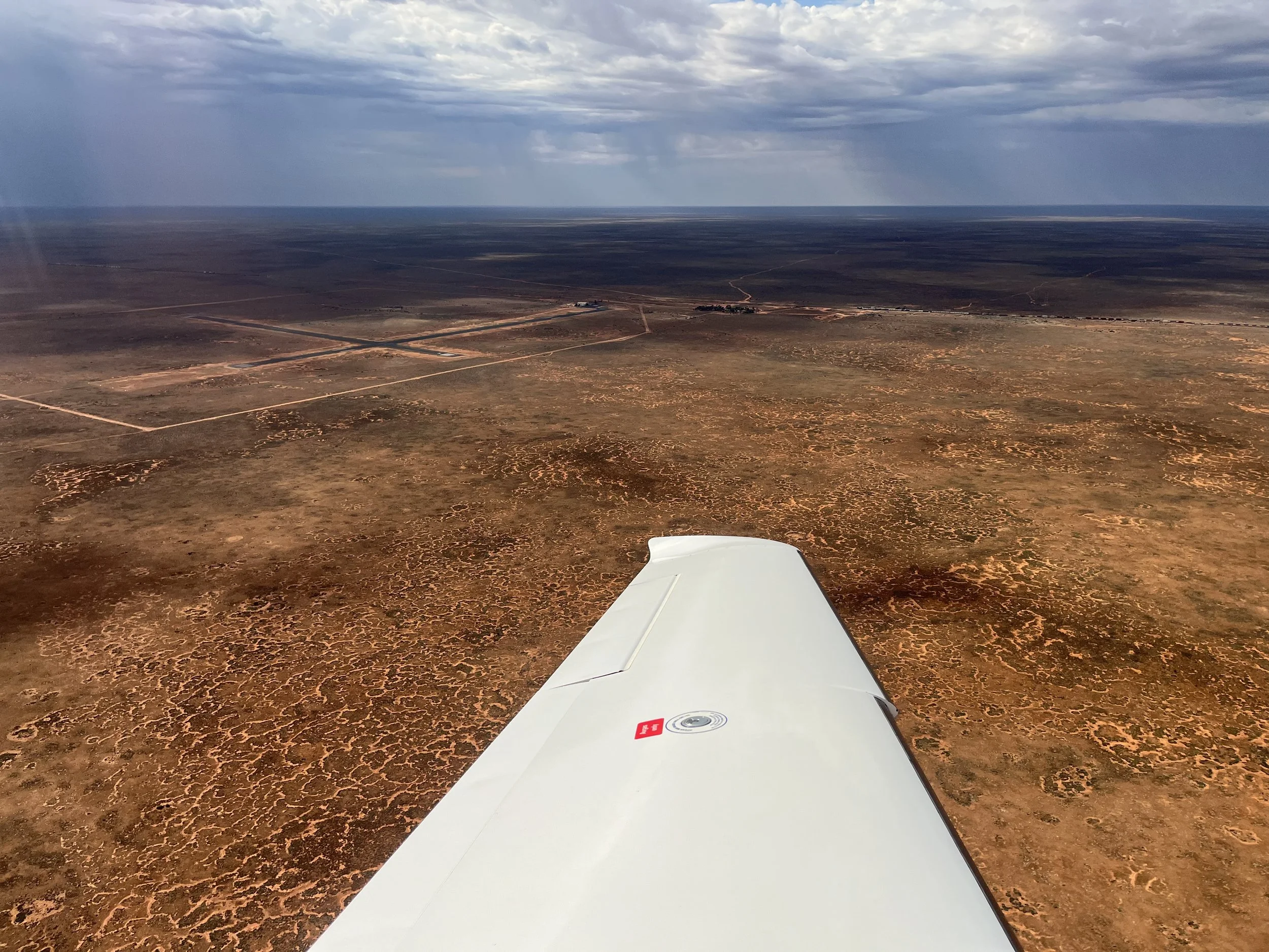 Photo taken from an airplane showing a vast desert landscape with reddish-brown terrain, some dirt roads, and a partly cloudy sky.