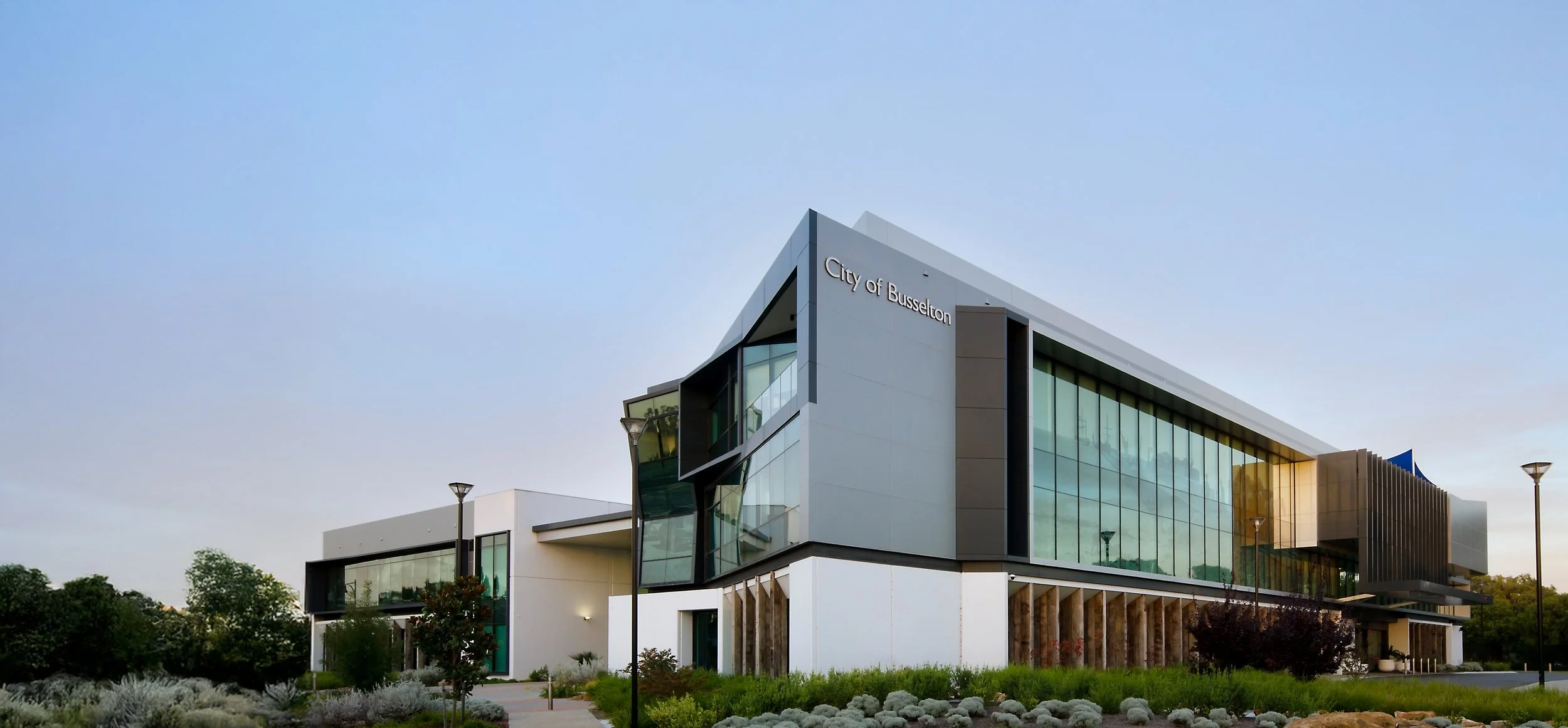 Modern building labeled 'City of Busselton' with large glass windows, surrounded by greenery, under a clear sky.