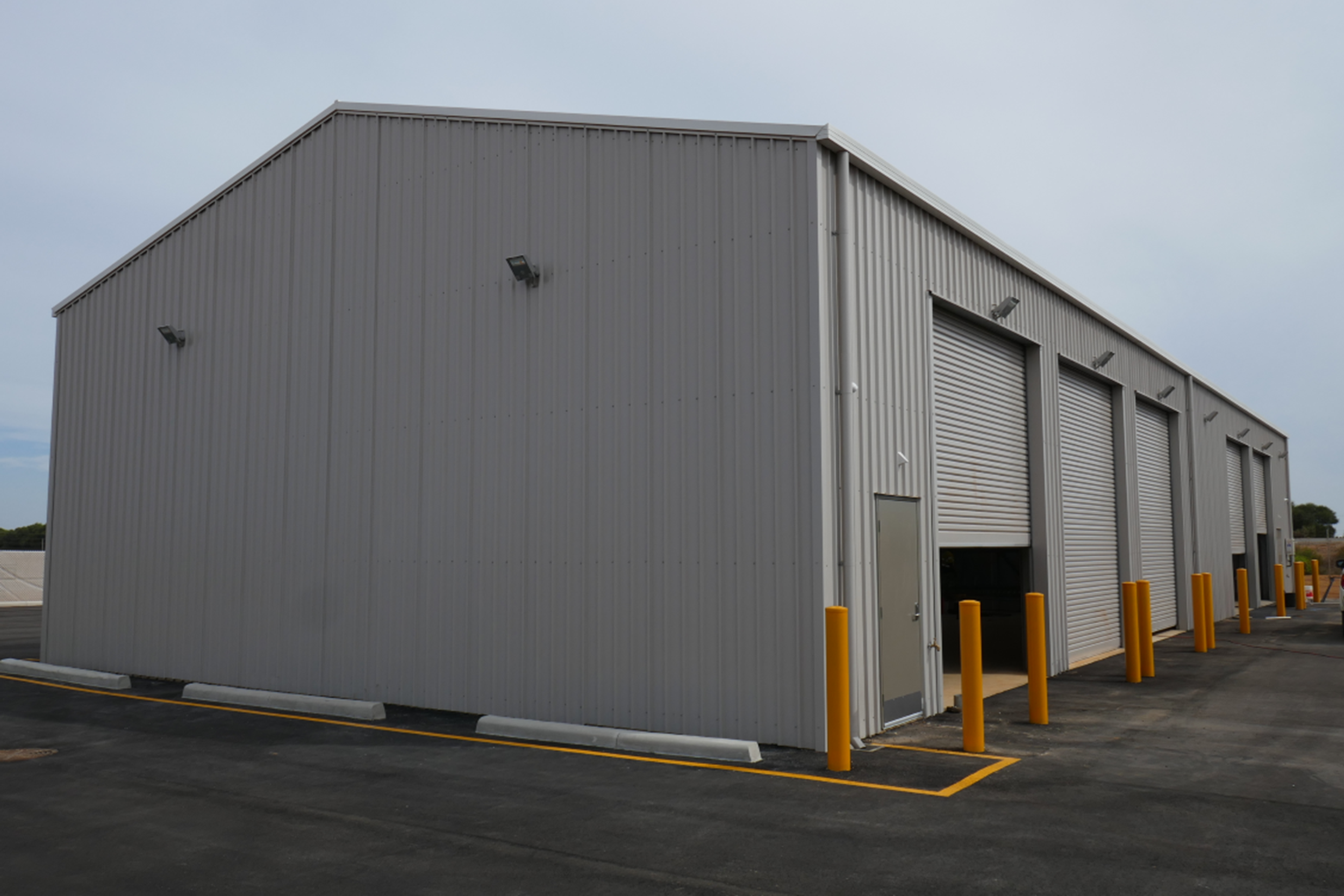 A large gray metal industrial warehouse with multiple roller doors and yellow safety bollards outside.