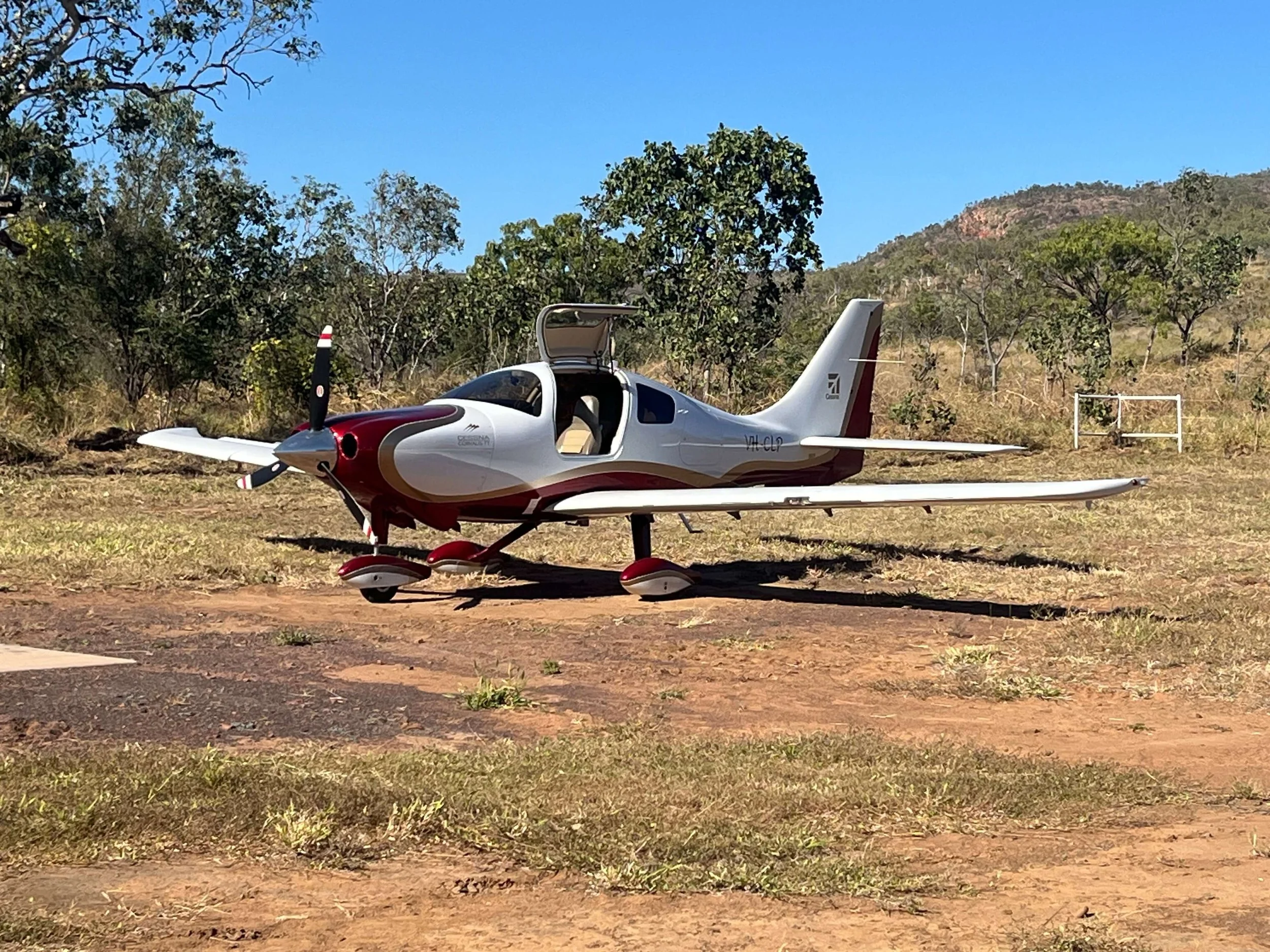 Small single-engine airplane parked on dirt ground with a background of trees, hills, and a clear blue sky.