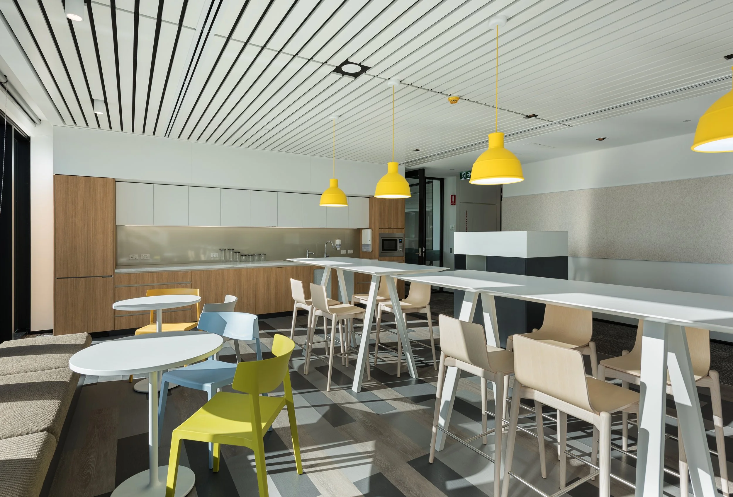 Modern kitchen and dining area with yellow pendant lights, various chairs, a long table, and a kitchenette in the background.
