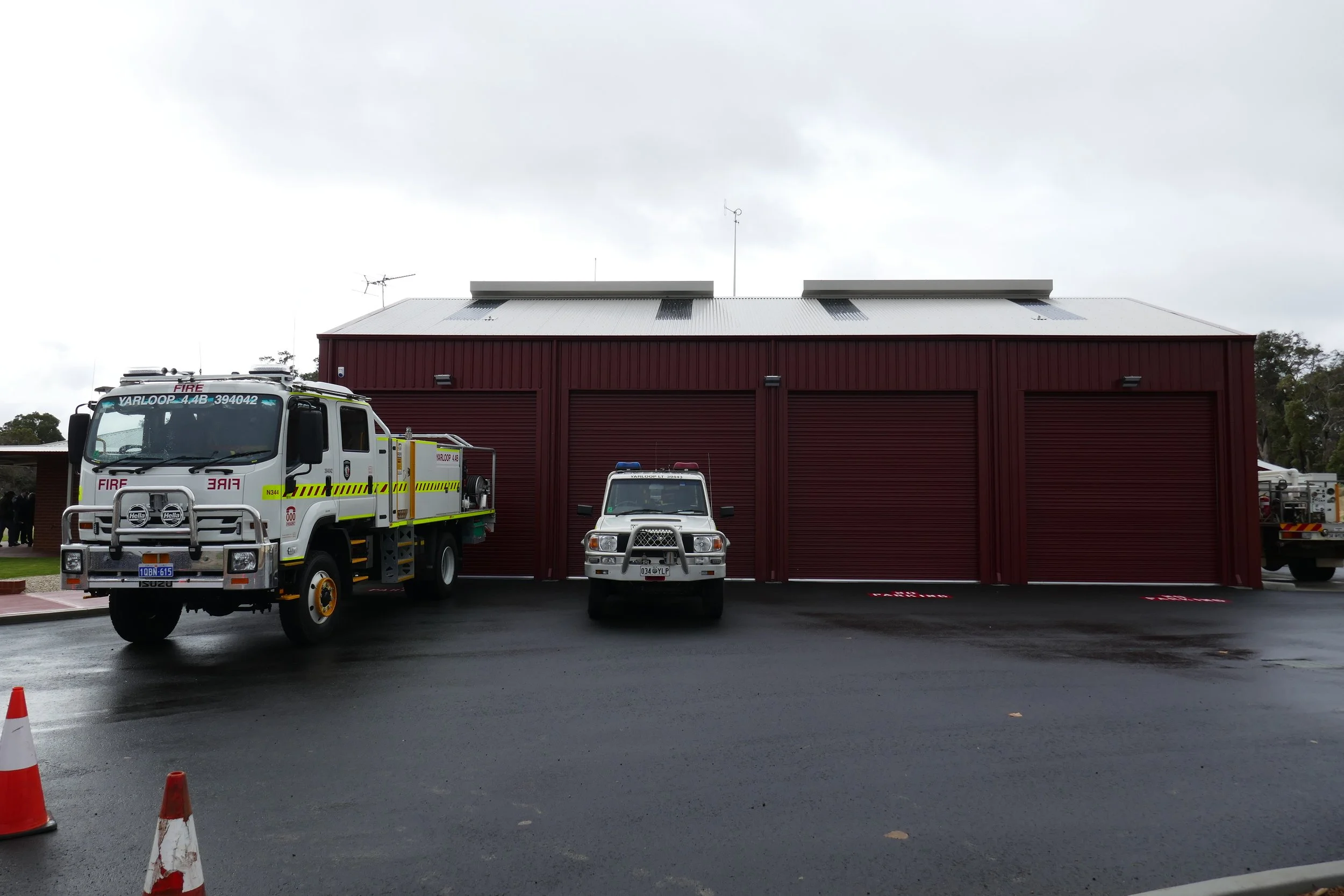 Three fire trucks parked in front of a red fire station on a wet pavement during overcast weather.