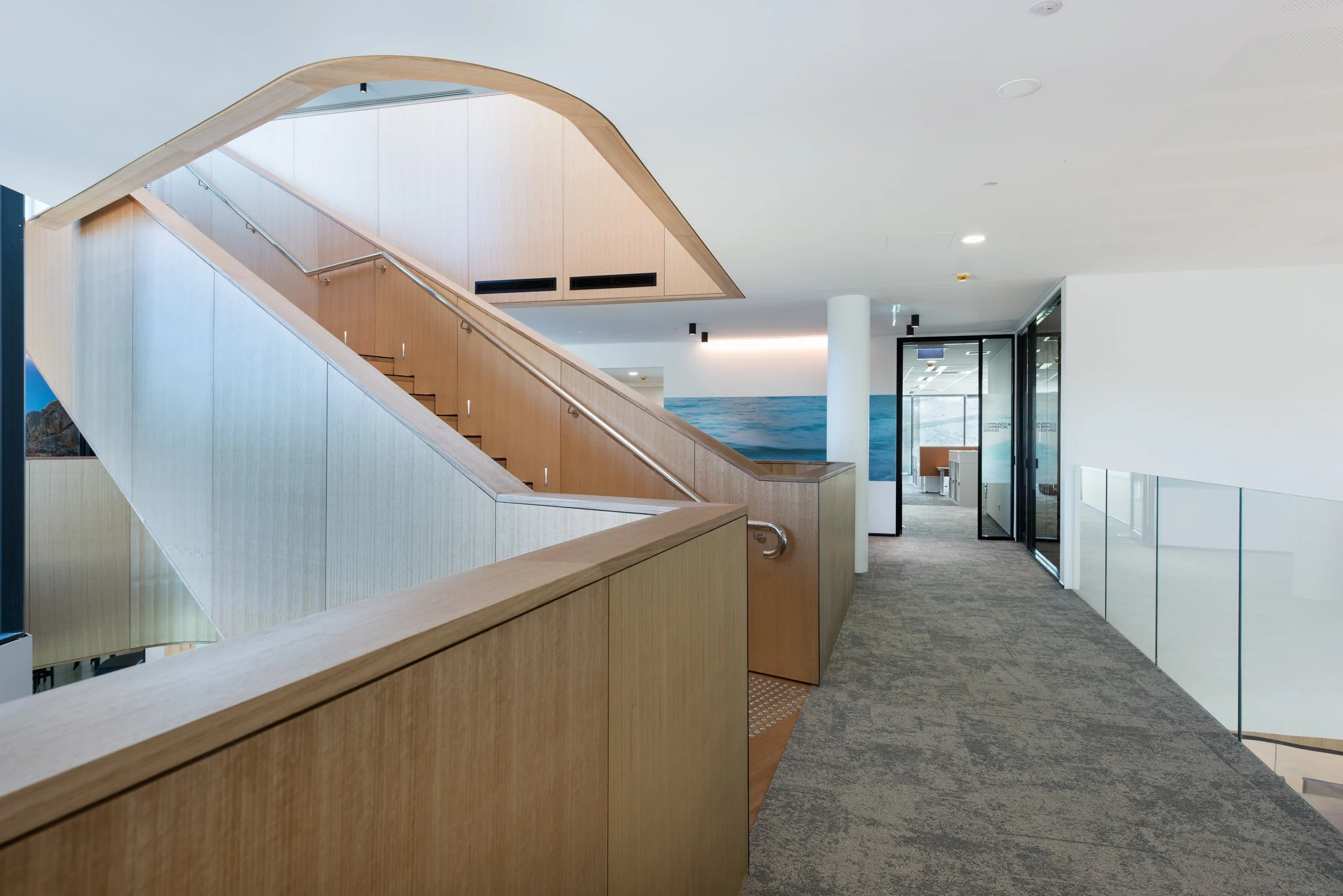 Interior of a modern building with a wooden staircase, glass walls, and a hallway with patterned carpet.