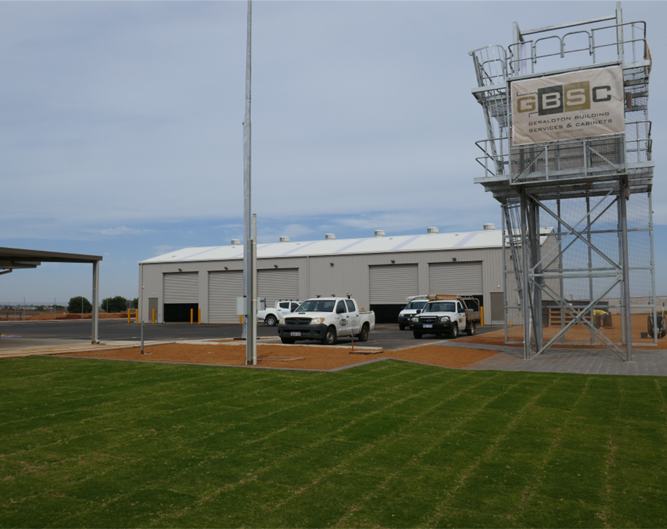 Industrial building with a large garage, several parked utility vehicles, and a metal watchtower with a banner reading "GBSC Geraldton Building Services & Cabinets" in a parking lot with a manicured lawn.