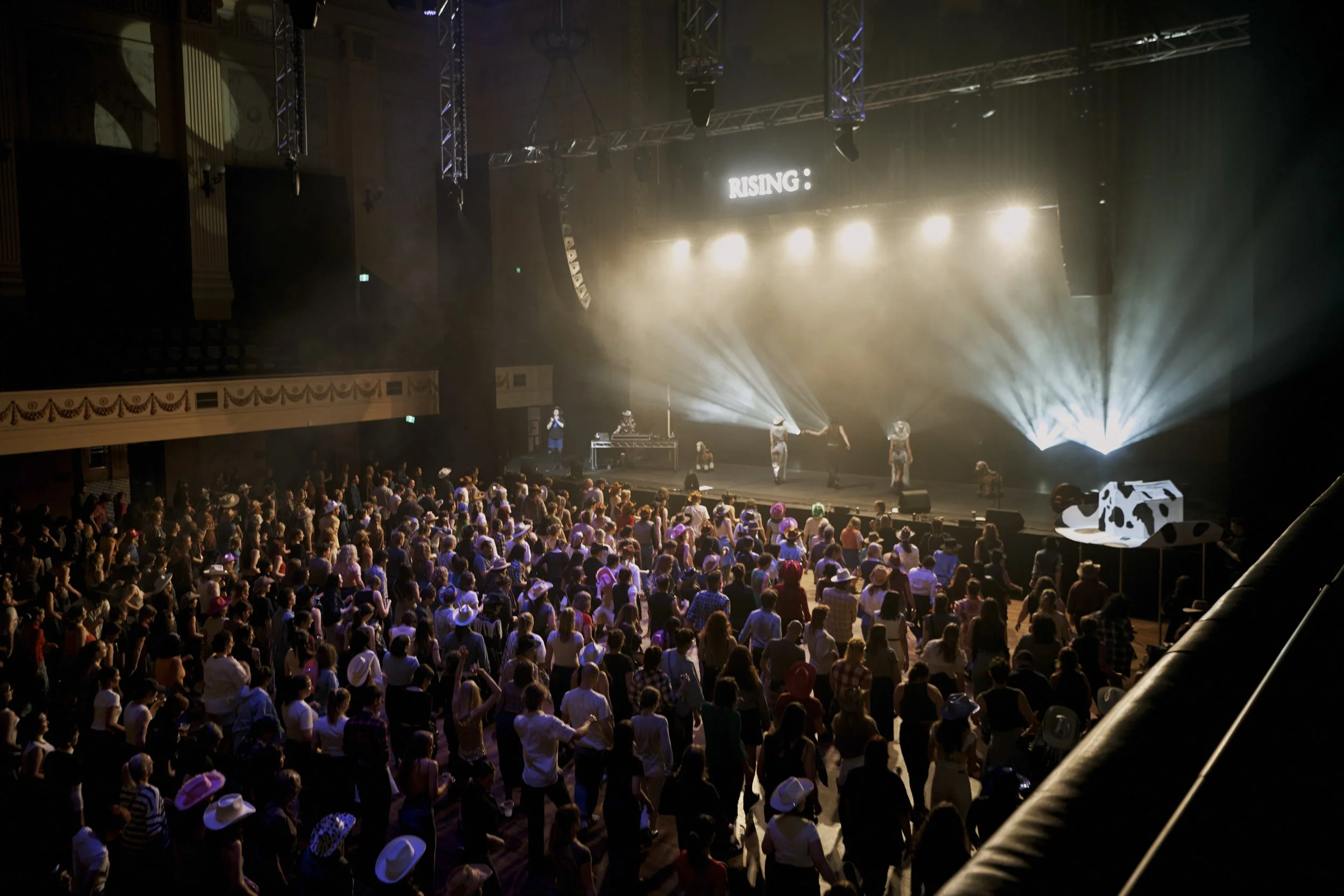 A concert or performance on stage with bright lights and performers, with a large audience watching in a theater or concert hall.