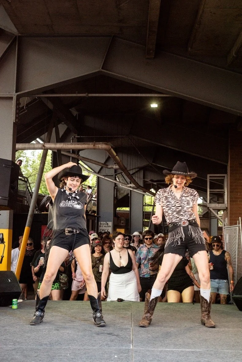 Two women in cowboy hats perform a dance on stage at an outdoor event, with a crowd watching behind them.