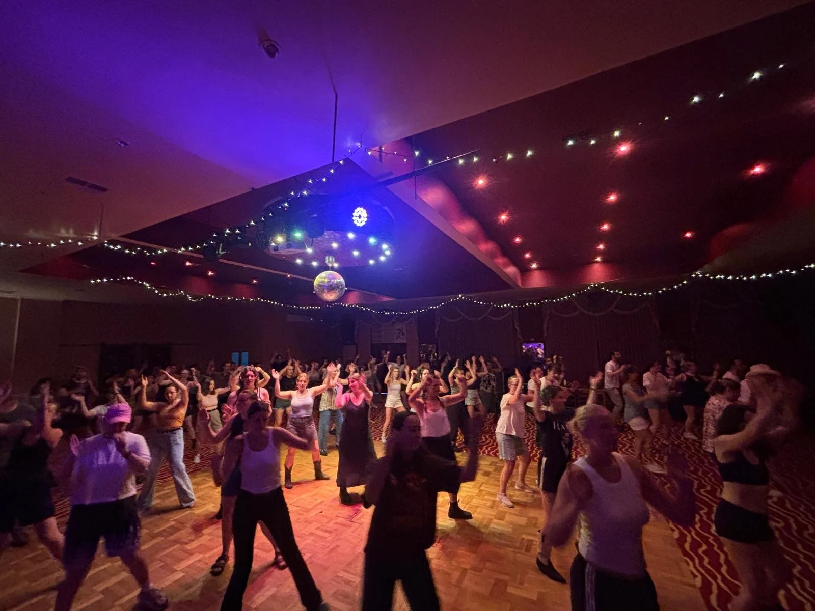 People dancing at a party in a dimly lit room with colorful lights and a disco ball.