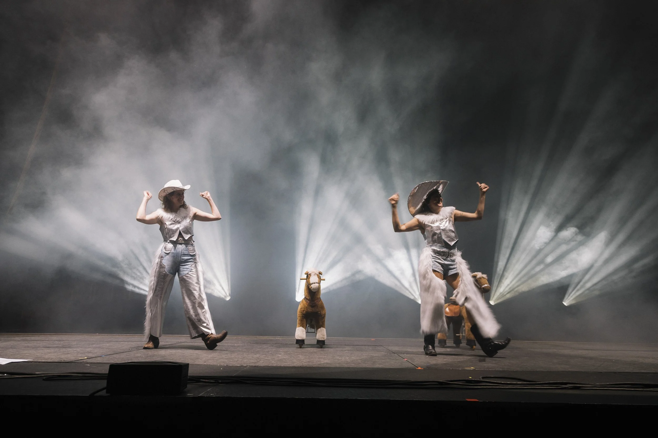 Two women wearing cowboy hats and furry pants dancing on a stage, with two small animals dressed as cows, in front of a smoky background and stage lights.
