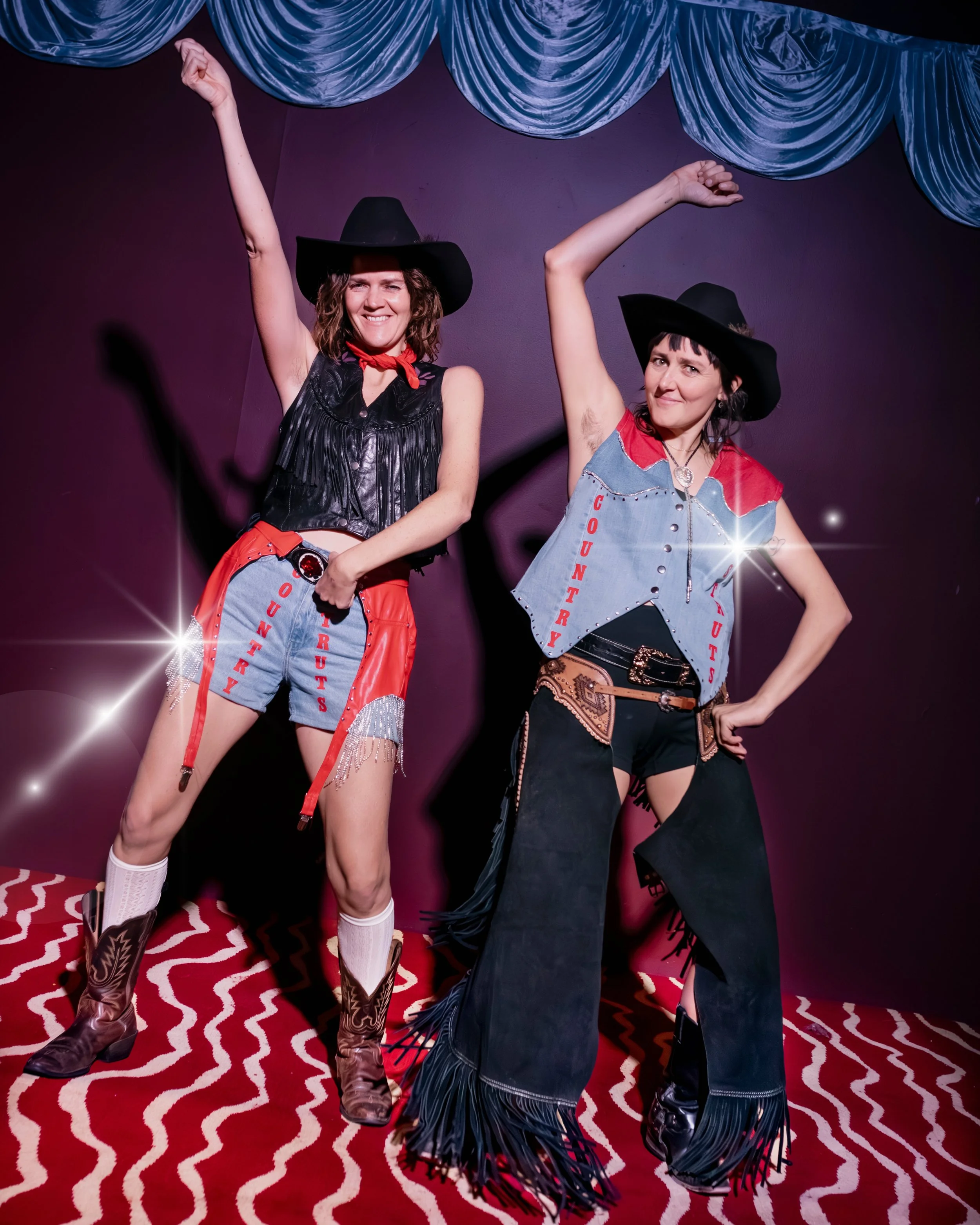Two women dressed in cowgirl costumes posing with arms raised, standing on a red patterned carpet in front of a purple wall with blue curtains.