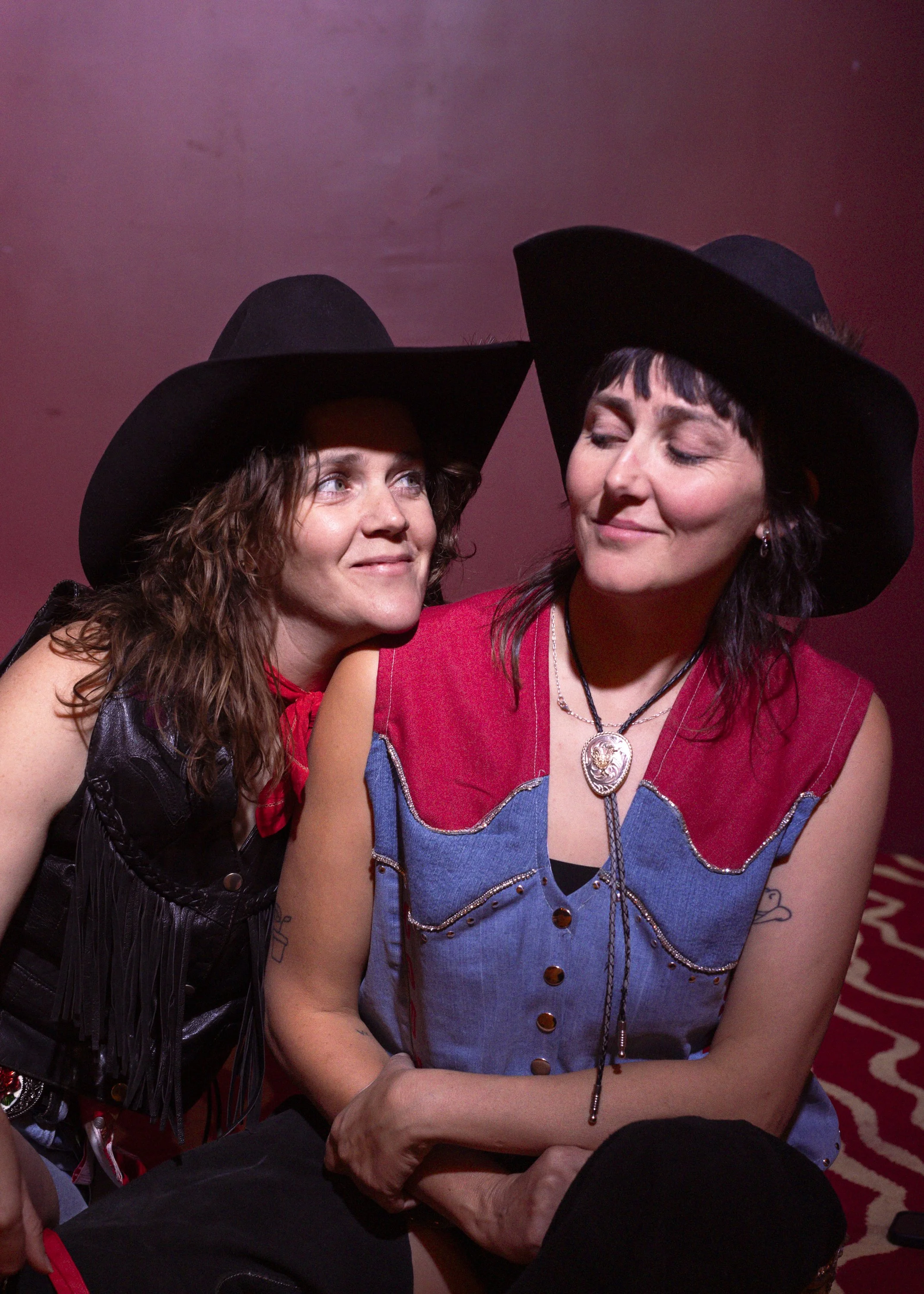 Two women wearing black cowboy hats, one with curly hair and the other with straight hair, sitting closely together, smiling, against a pinkish background.