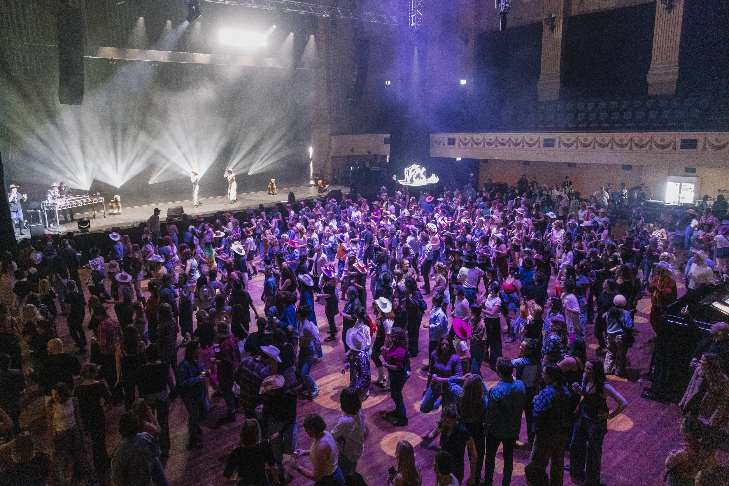 Crowd of people dancing in a large ballroom during a country-western dance event, with some wearing cowboy hats and western attire, stage with performers and purple lighting in the background.