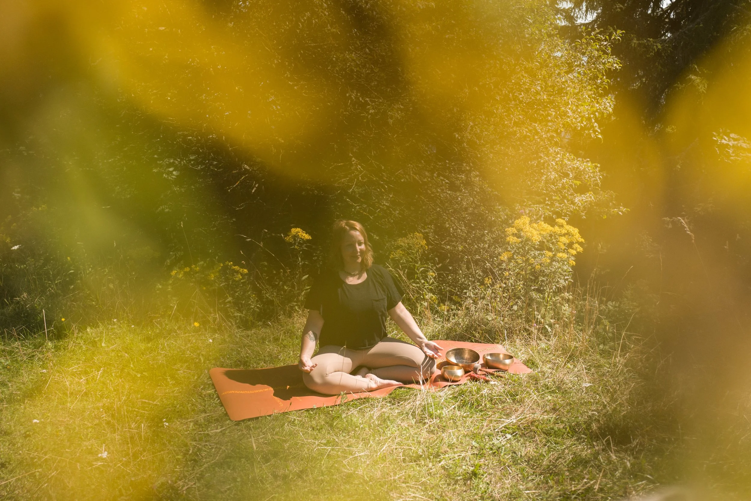Eine Frau sitzt auf einer Yoga-Matte im Freien, umgeben von Büschen und Bäumen, bei schönem Wetter, mit mehreren Klangschalen vor ihr, während sie entspannt meditiert.