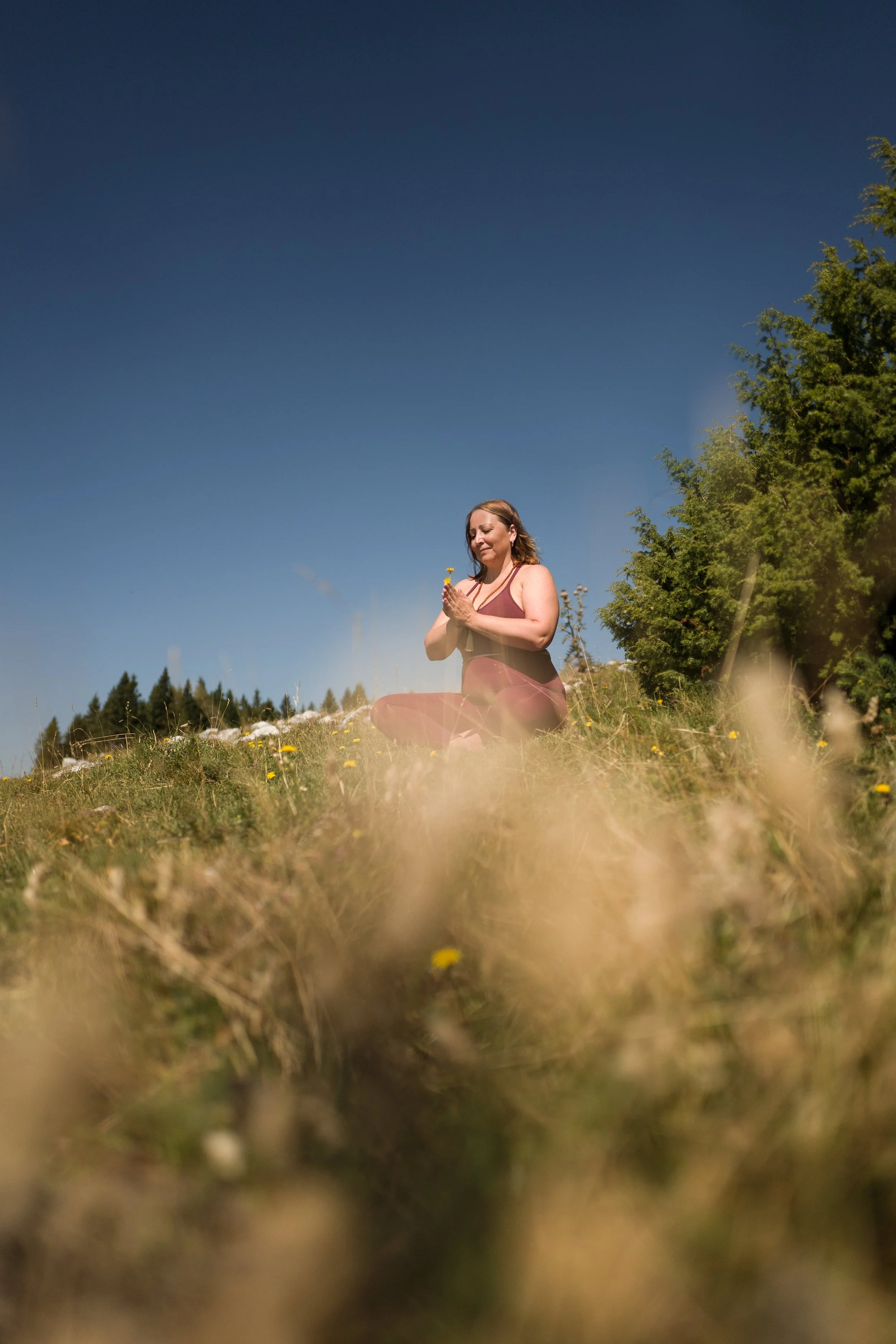 Frau meditiert im Freien auf einer Wiese bei sonnigem Wetter, umgeben von Gras und Bäumen.