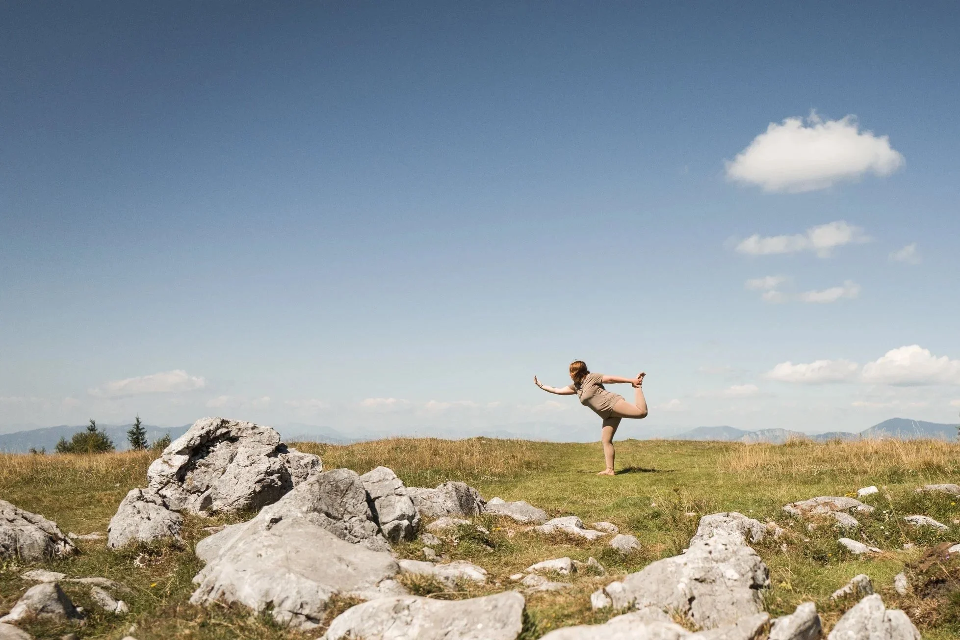 Eine Person praktiziert Yoga im Freien auf einer Wiese mit Felsen, im Hintergrund Berge und ein blauer Himmel mit wenigen Wolken.