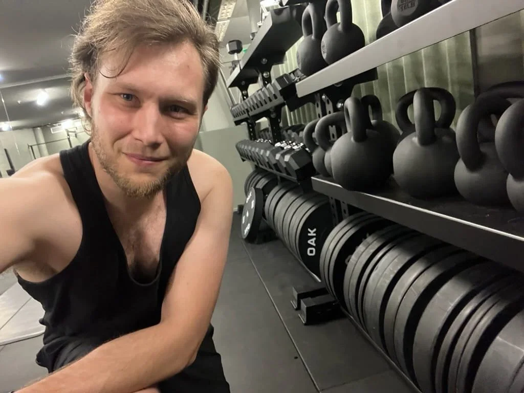 man står i ett gym vid ett viktställ kettlebells och viktplattor och tittar in i kameran
