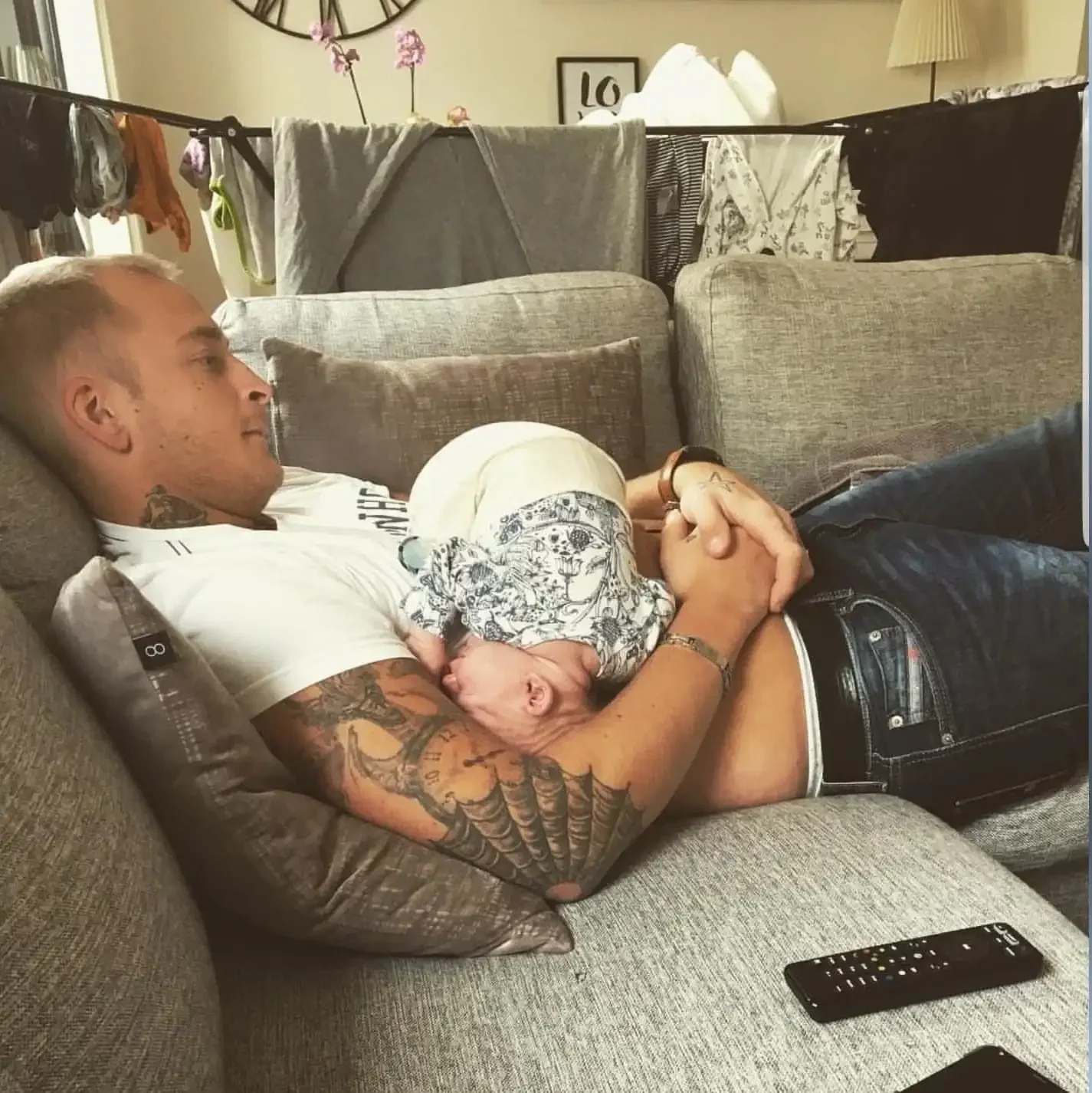 En man vilar i en soffa med tatueringar på armen, håller en bebis klädd i en mönstrad outfit, med en kudde bakom huvudet och en TV-fjärrkontroll på soffan intill.