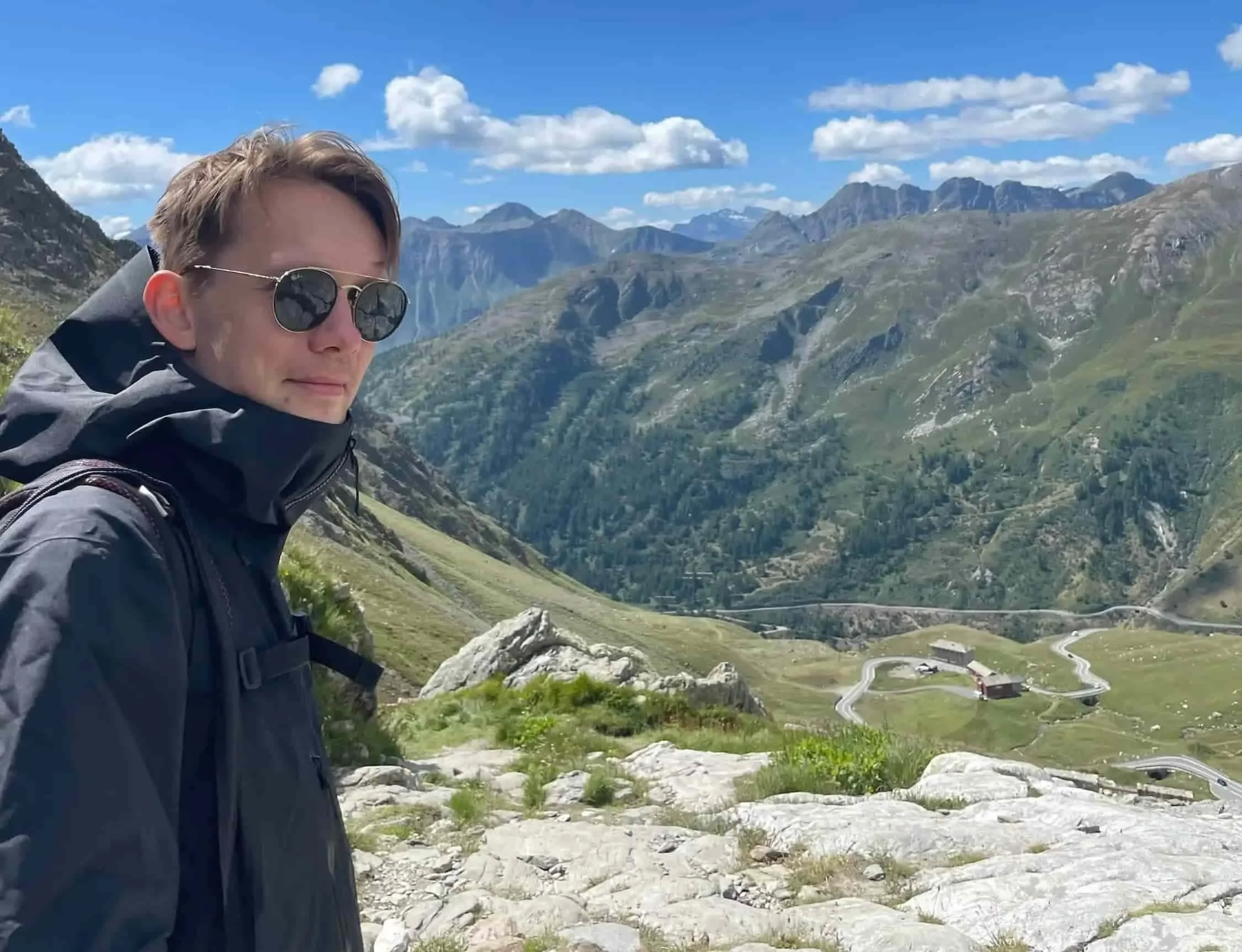 En man med mörk jacka tittar in i kameran med öppna landskap och blå himmel bakom sig.jpg
